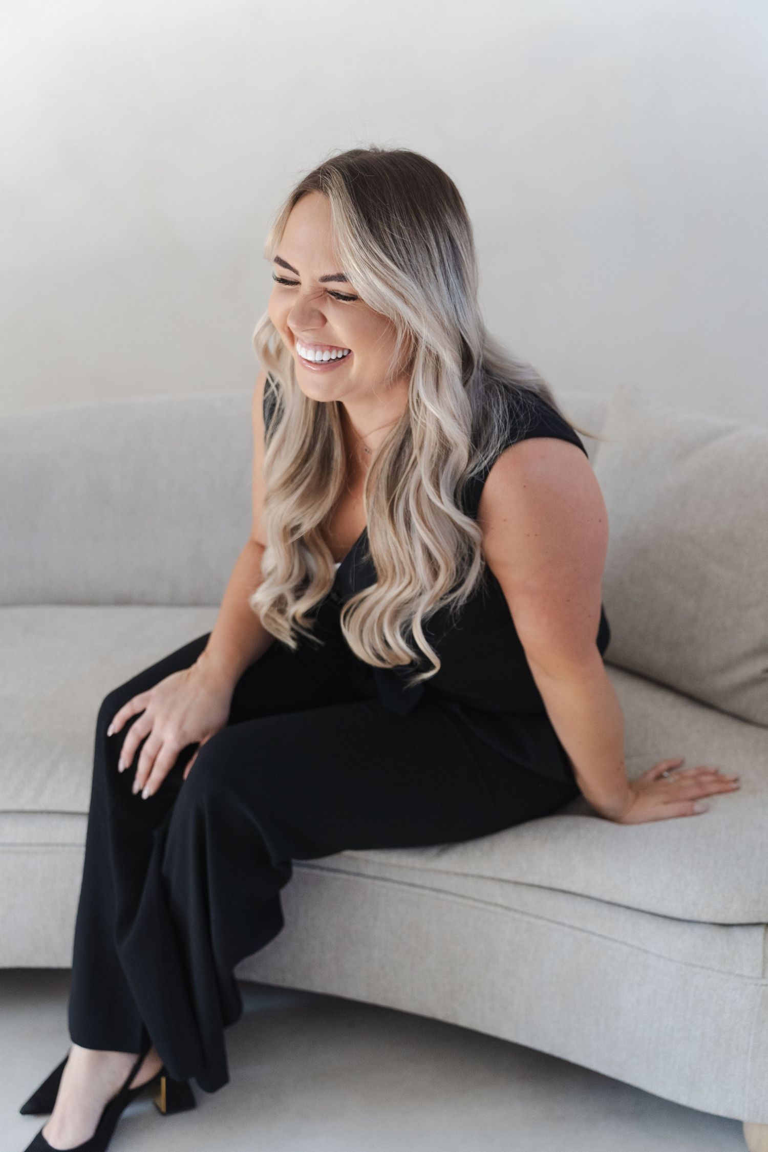 A woman with platinum blonde hair wearing a black dress lounges elegantly across a light gray modern sofa.