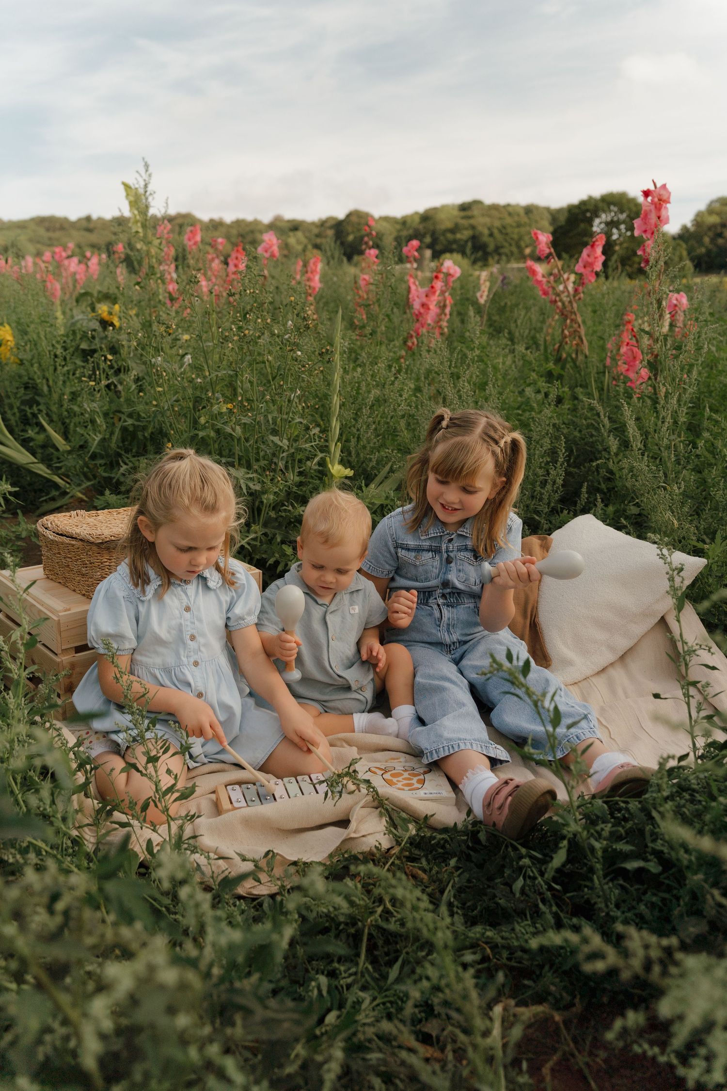 Three young children sit on a blanket in a wildflower meadow, wearing blue outfits, enjoying a sunny outdoor picnic.