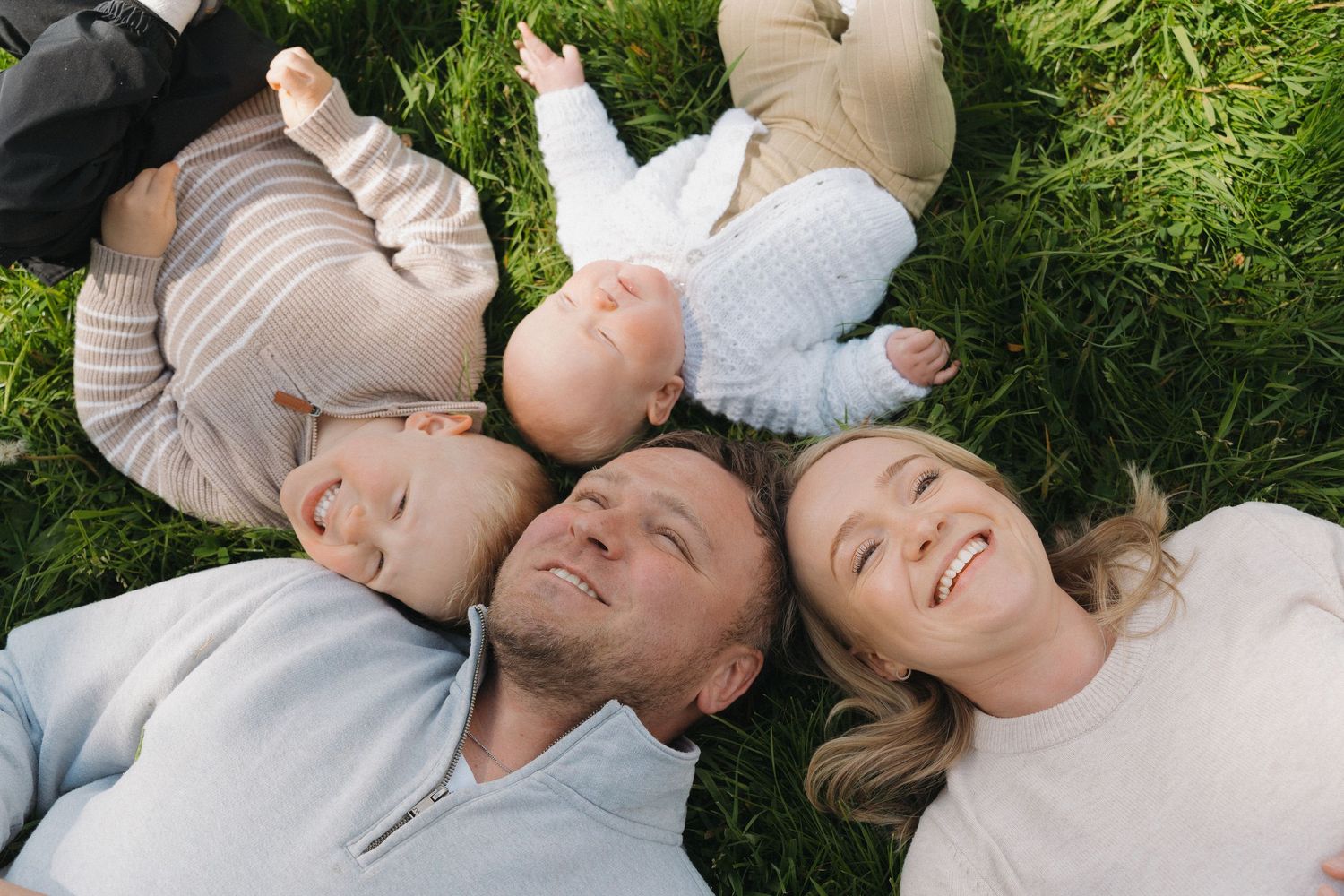 Happy family of four lying on green grass outdoors, smiling and enjoying a sunny day together.