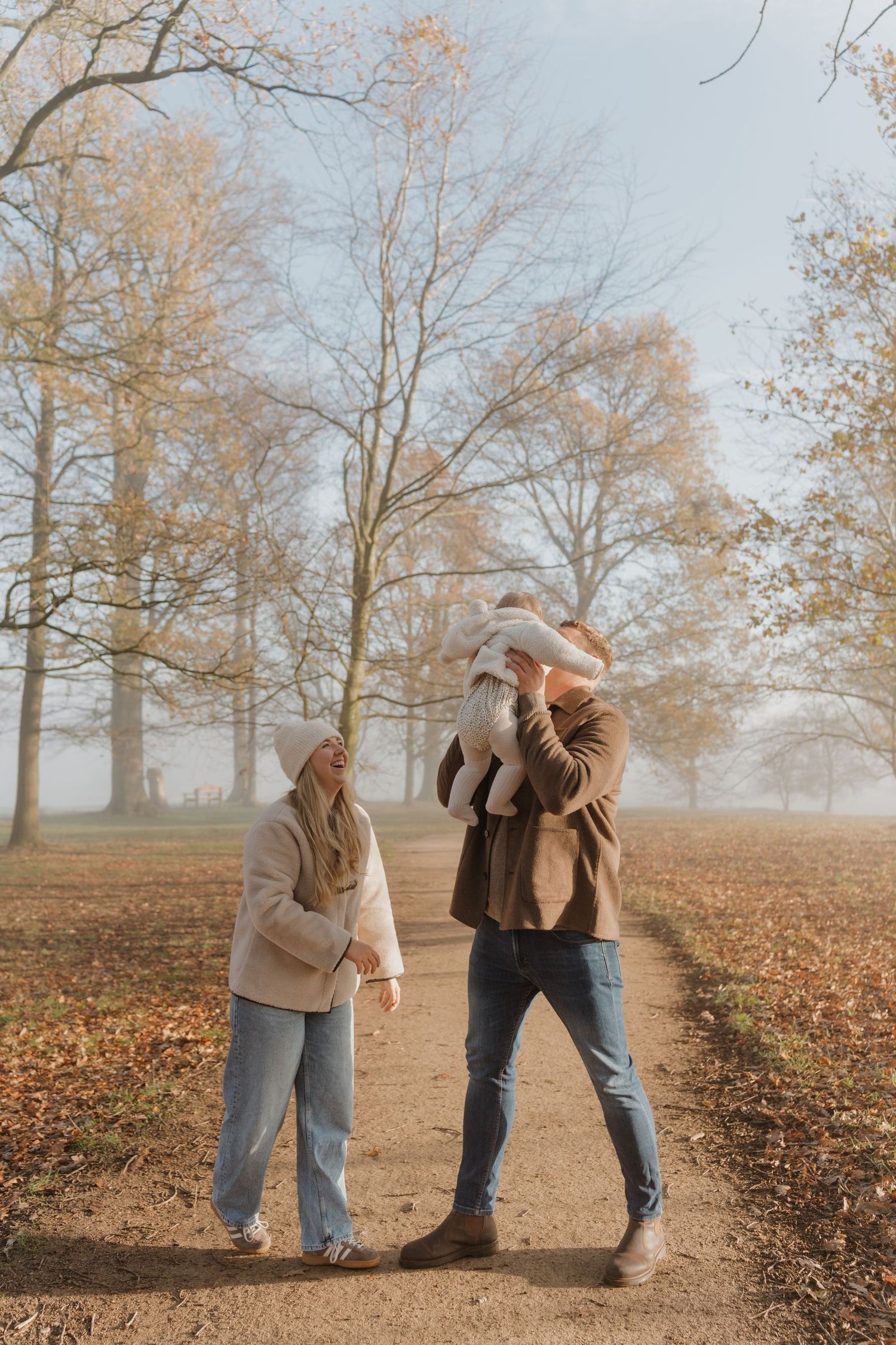 A couple and their child enjoy an autumn walk in a misty park with bare trees and fallen leaves.