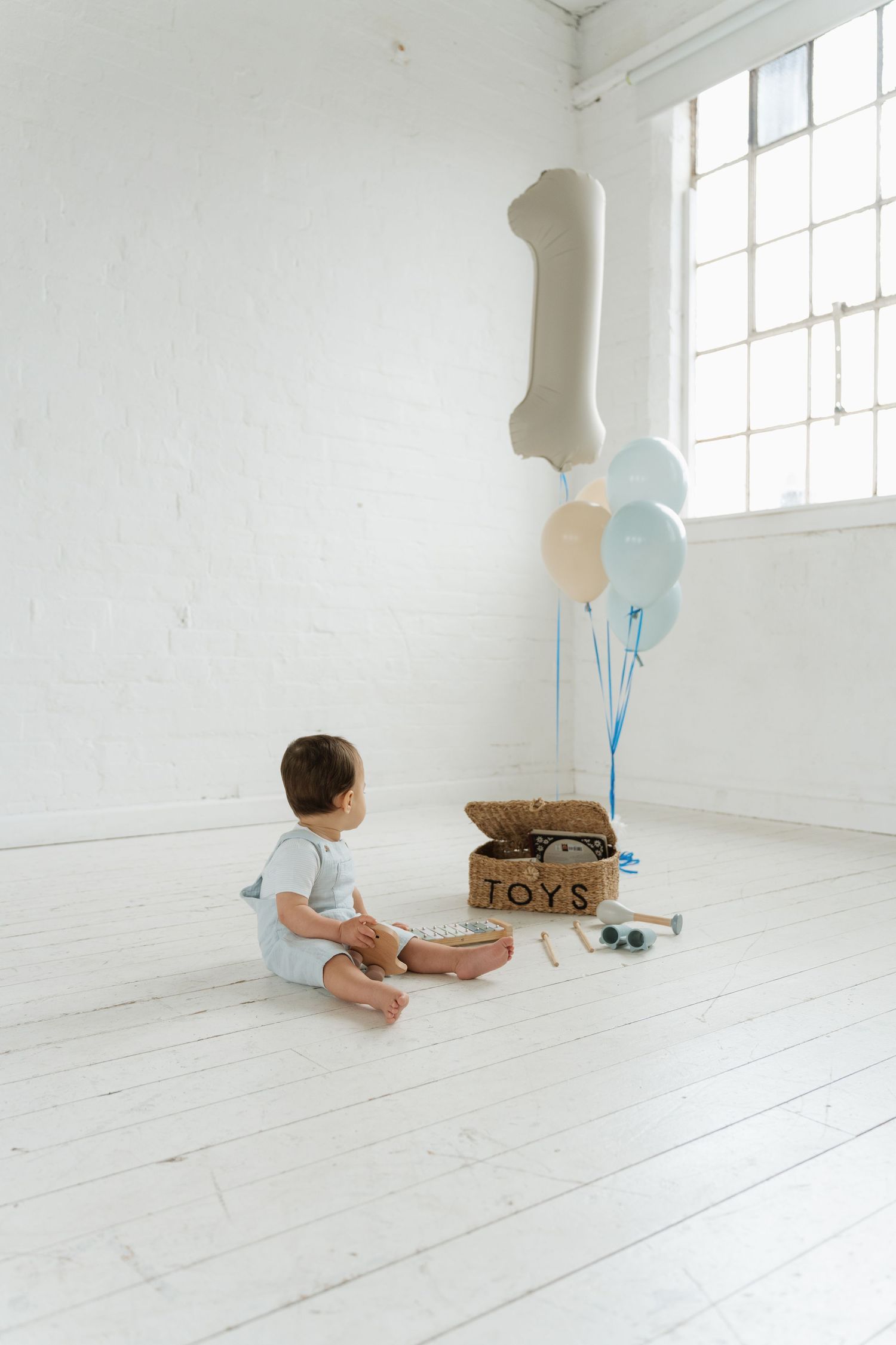 Baby boy sits on white floor with toys box and number one balloons celebrating his first birthday.