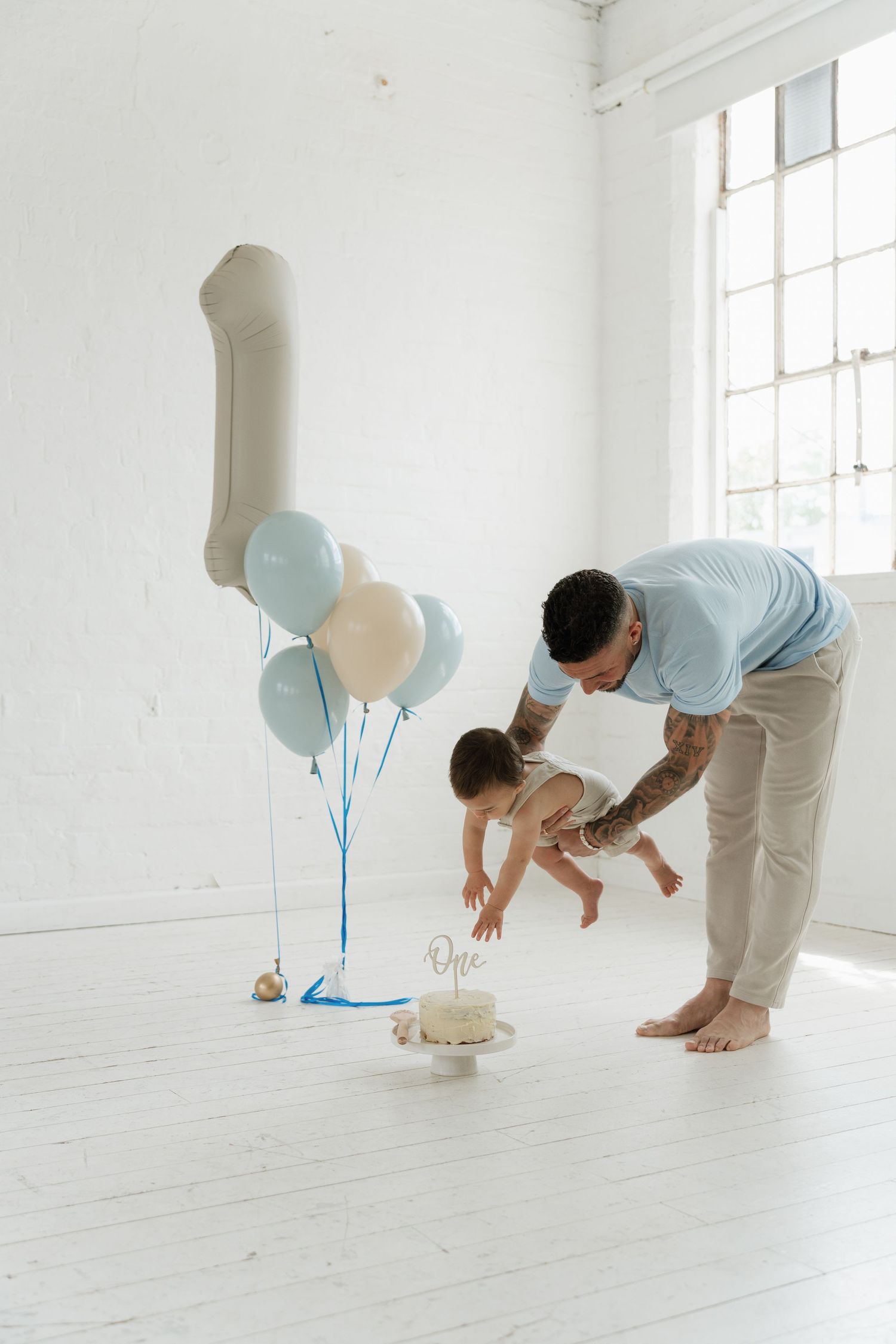 Father helping baby touch first birthday cake surrounded by blue and white balloons in bright white room.