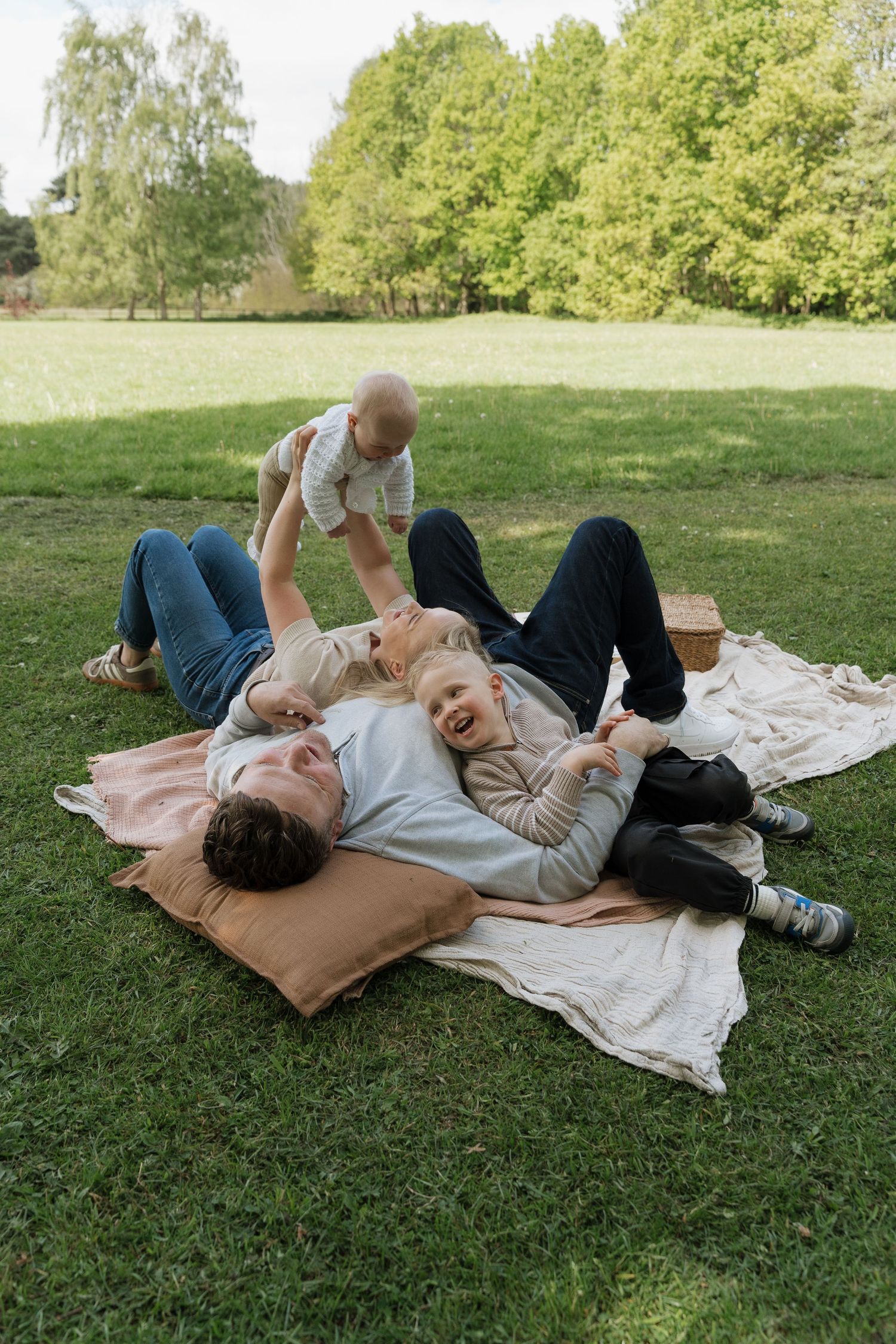 Family enjoying a sunny picnic on a blanket in a green park, with parents laughing and playing with two young children.