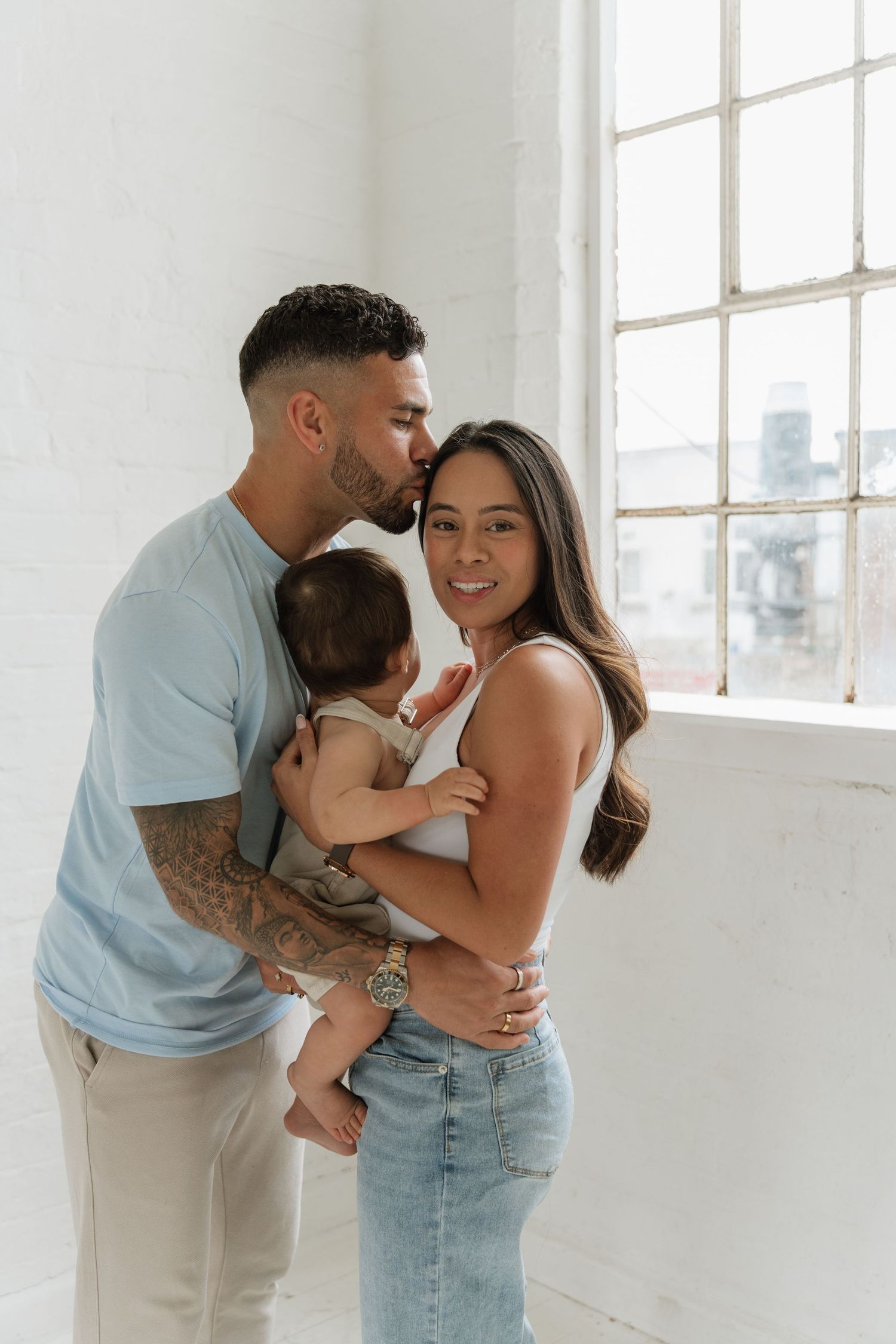 Young parents share a tender moment kissing their newborn baby by a bright window in a white brick room.