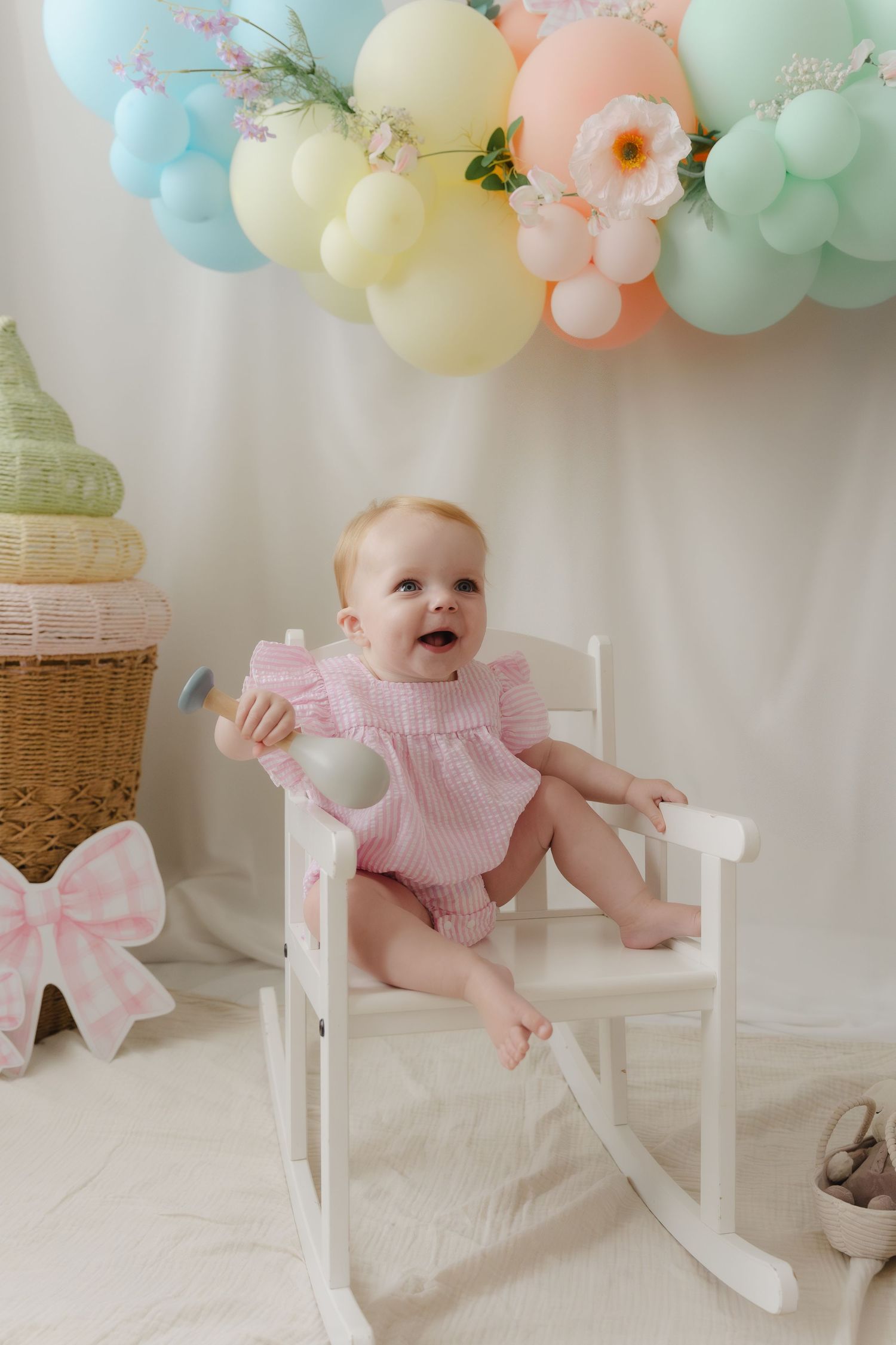 A baby in a pink dress sits in a white rocking chair under pastel balloon decorations.