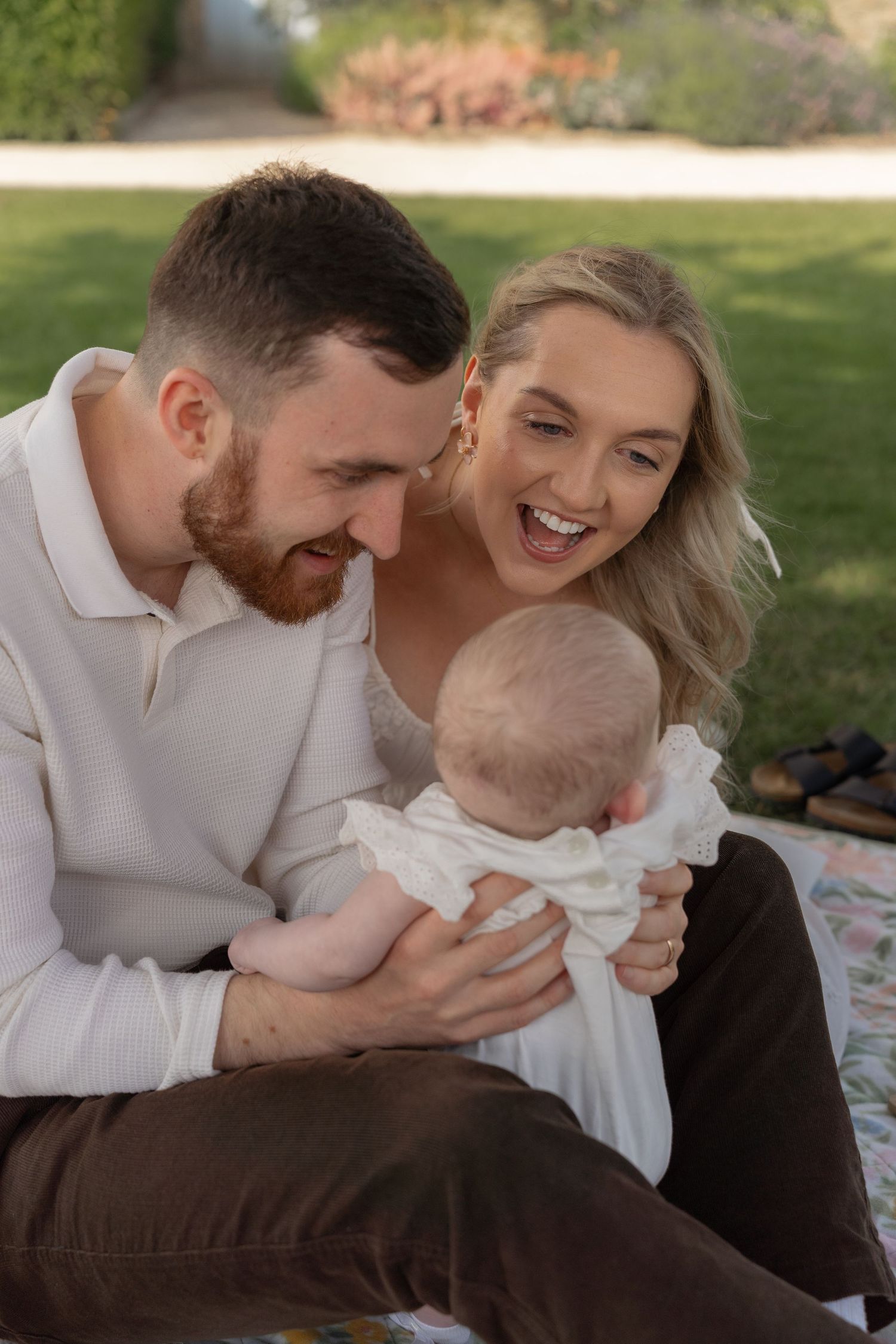 A couple shares joyful moments with their baby during an outdoor photo session in a sunlit garden setting.
