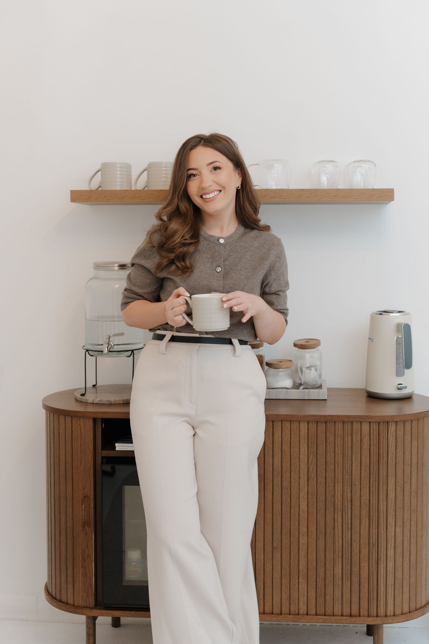 Smiling woman in beige outfit holding a mug leaning against a wooden coffee bar with shelves.