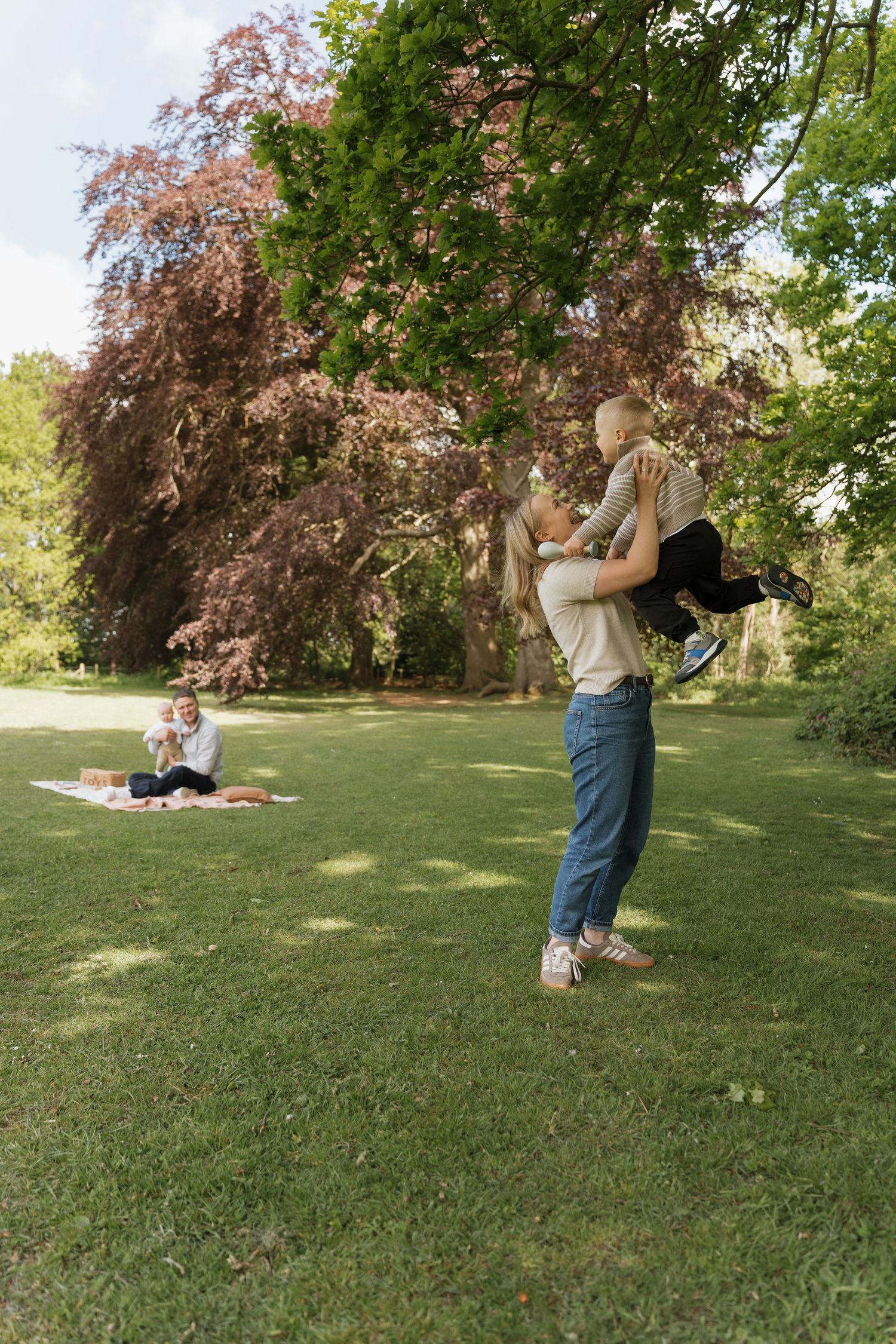 Family enjoying a sunny park picnic, with a parent lifting a child joyfully while another adult relaxes on a blanket.