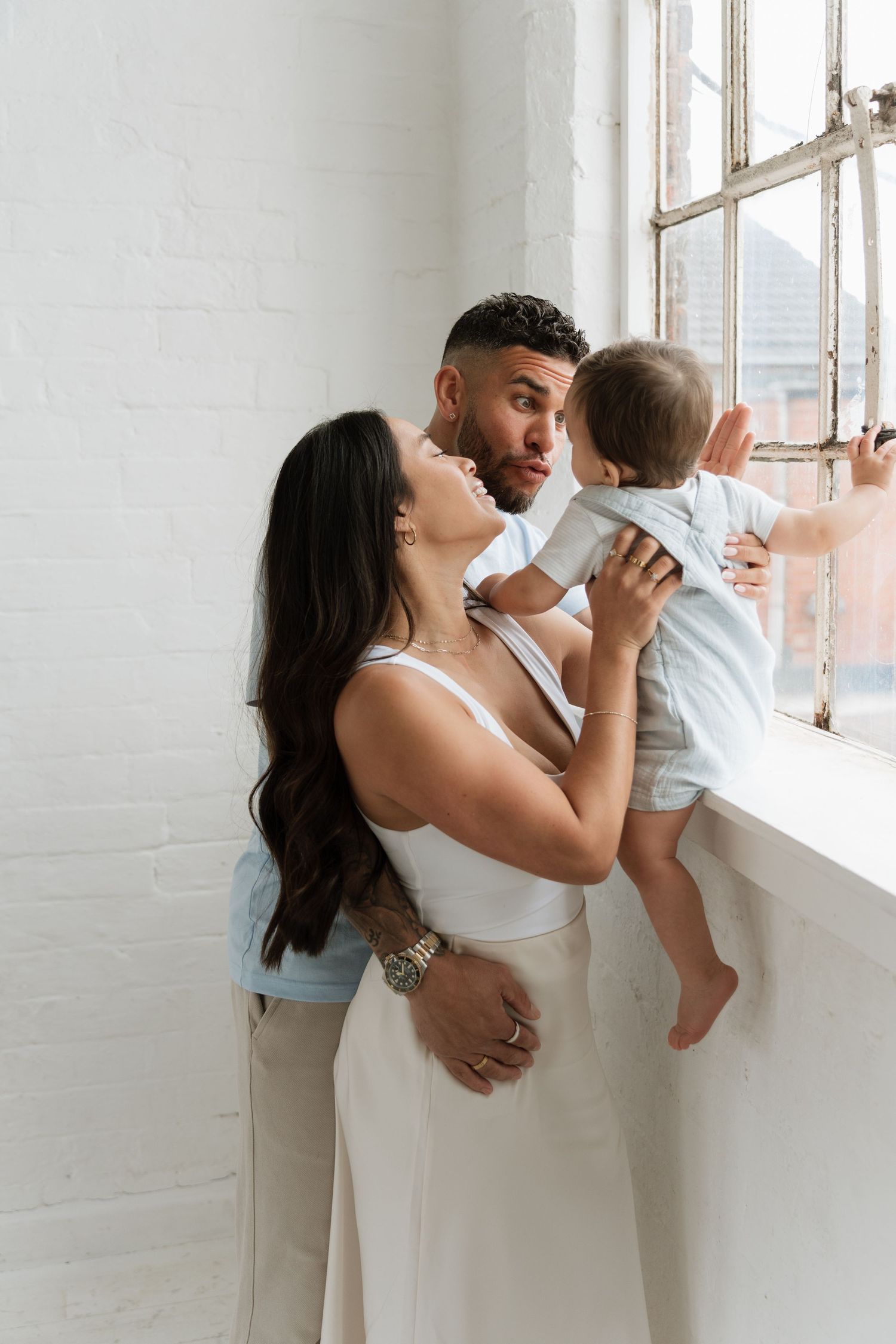 A couple shares a tender moment with their baby by a window in a bright white brick room.