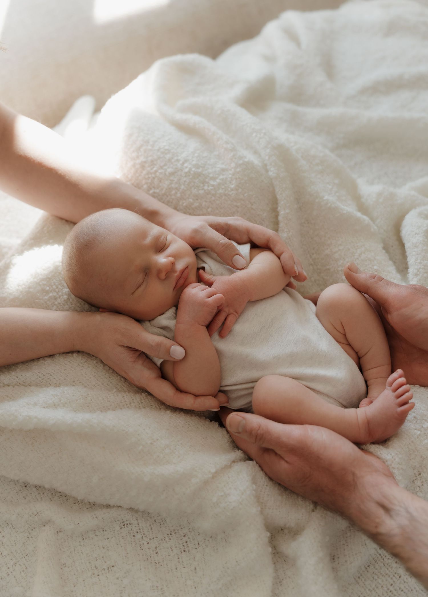 Newborn baby sleeping peacefully on white blanket while parents gently cradle with loving hands. Home Newborn Photography Derbyshire.