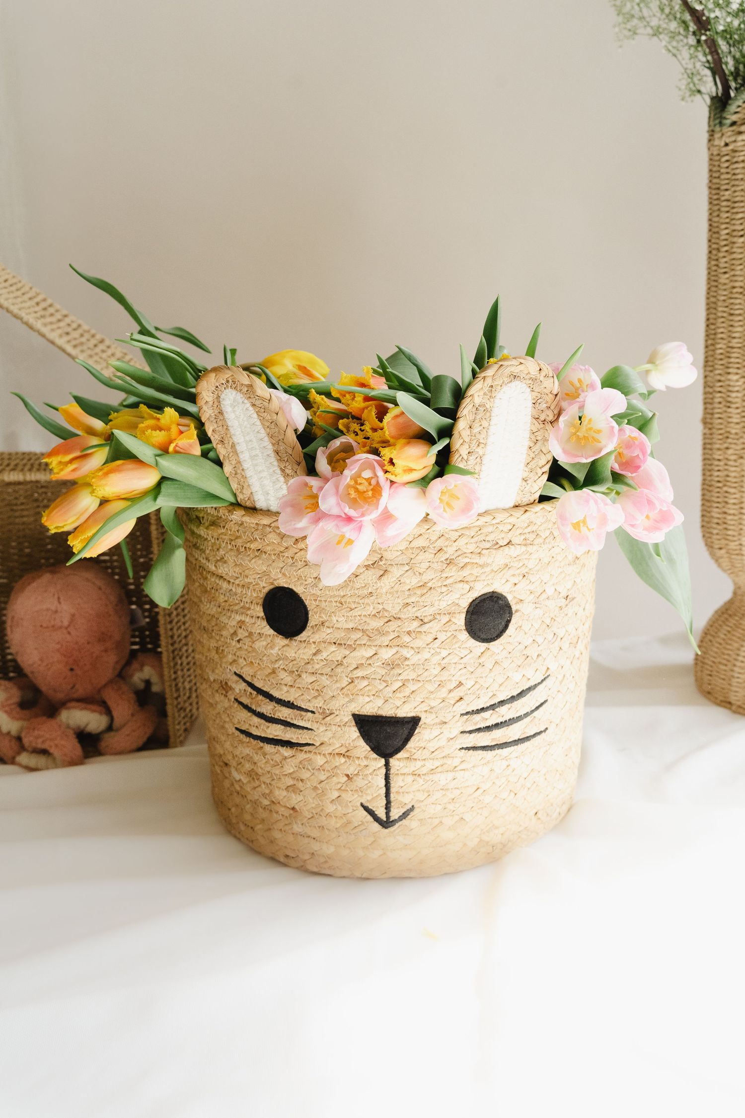 Straw basket with cat face design decorated with pink and orange flowers and greenery on white surface.