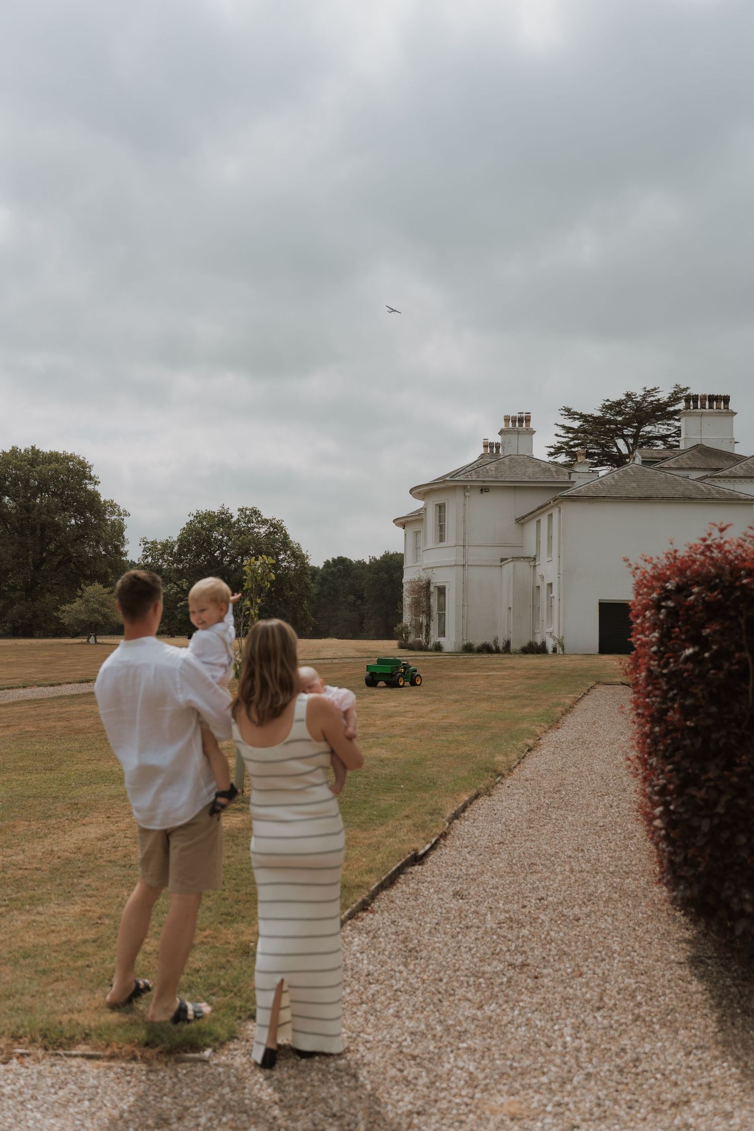 A family of three walks toward a grand white country house on a gravel path, with lush green lawns under a cloudy sky.