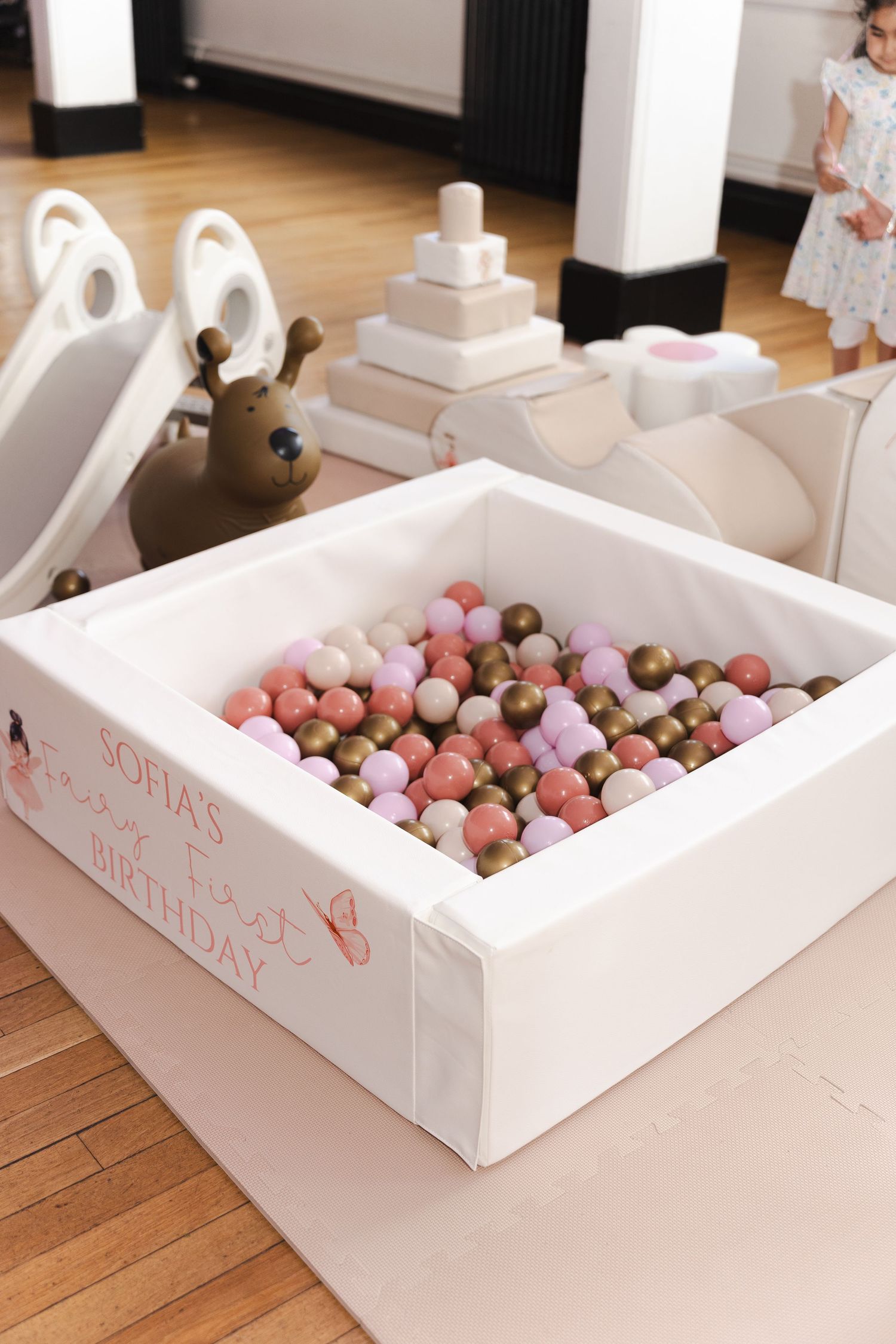 Pink, gold, and white ball pit for Sofia's first birthday party with toys and a slide in the background.