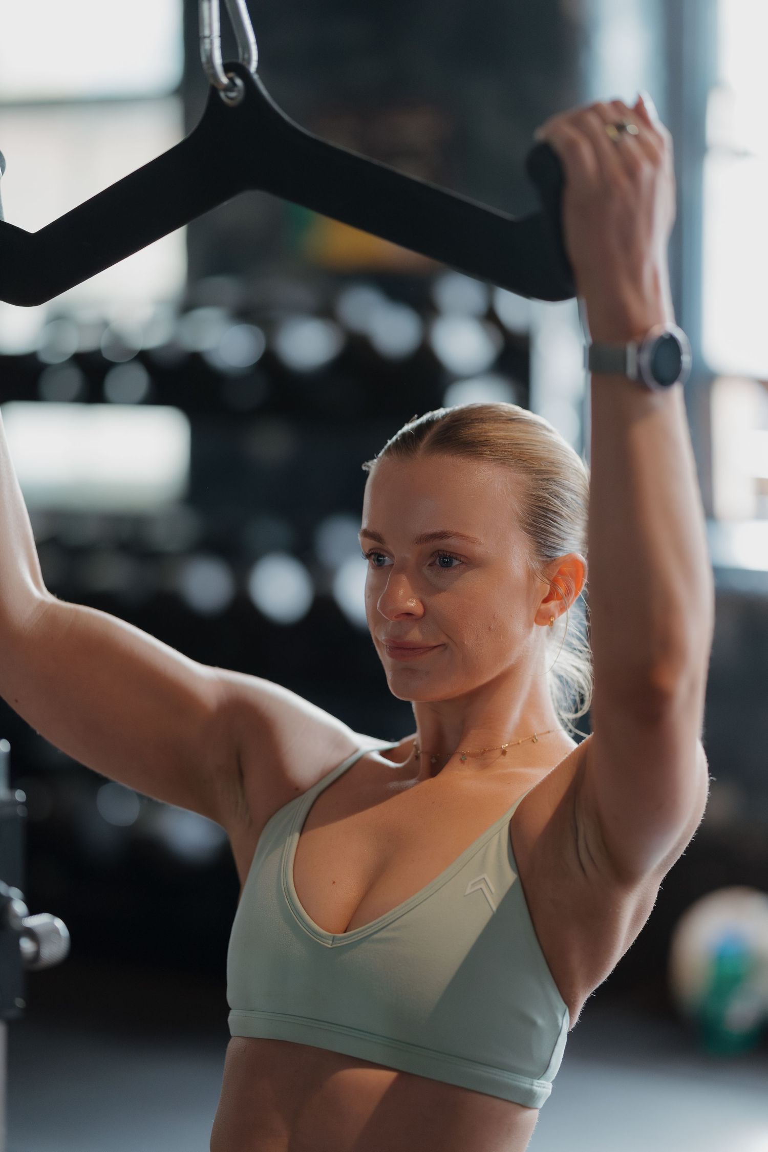 Fit woman in sports bra gripping gym bar overhead, showcasing toned arms and athletic physique in modern fitness center.