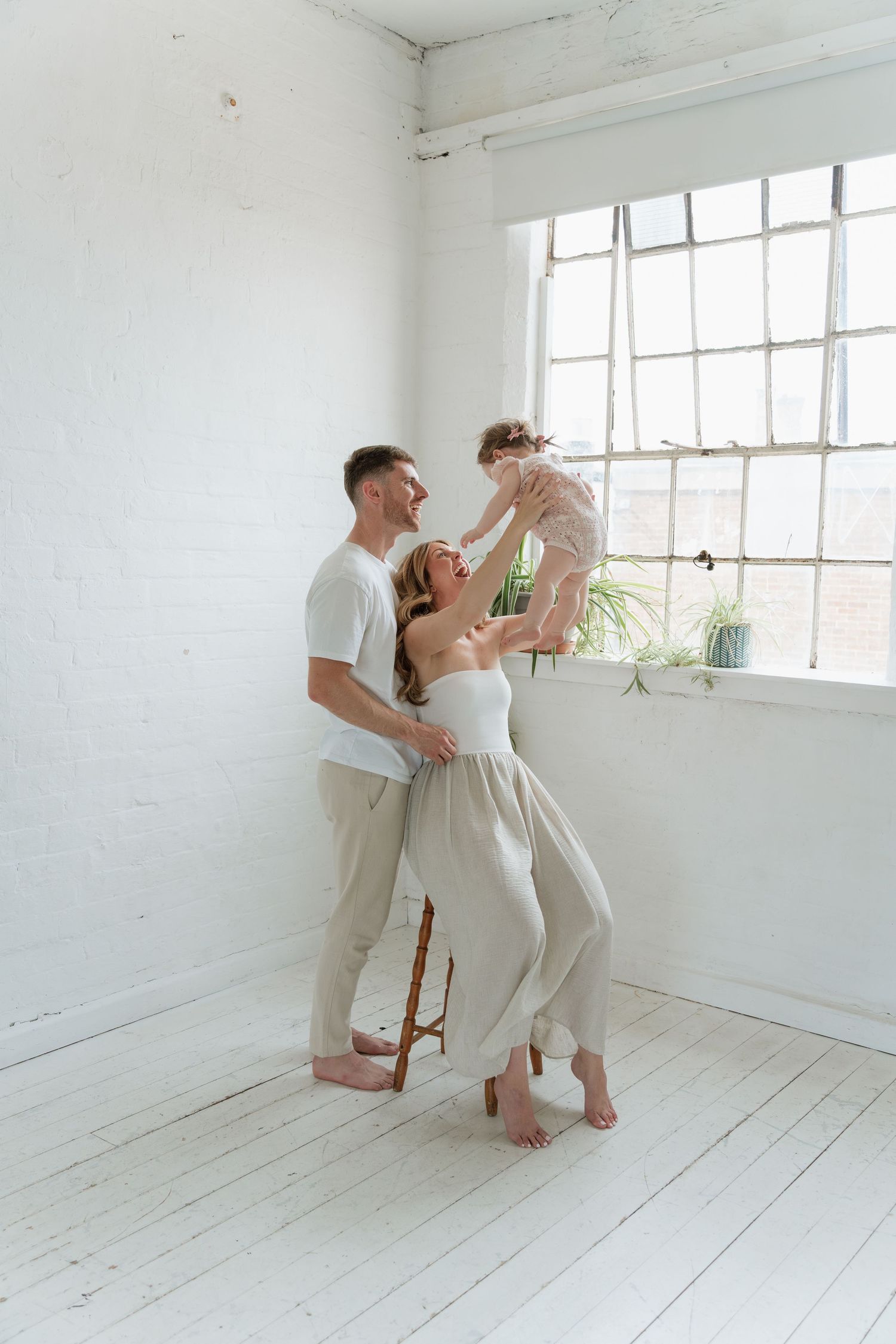 A joyful family in neutral tones plays together in a bright white room, with parents lifting their toddler near a large window.