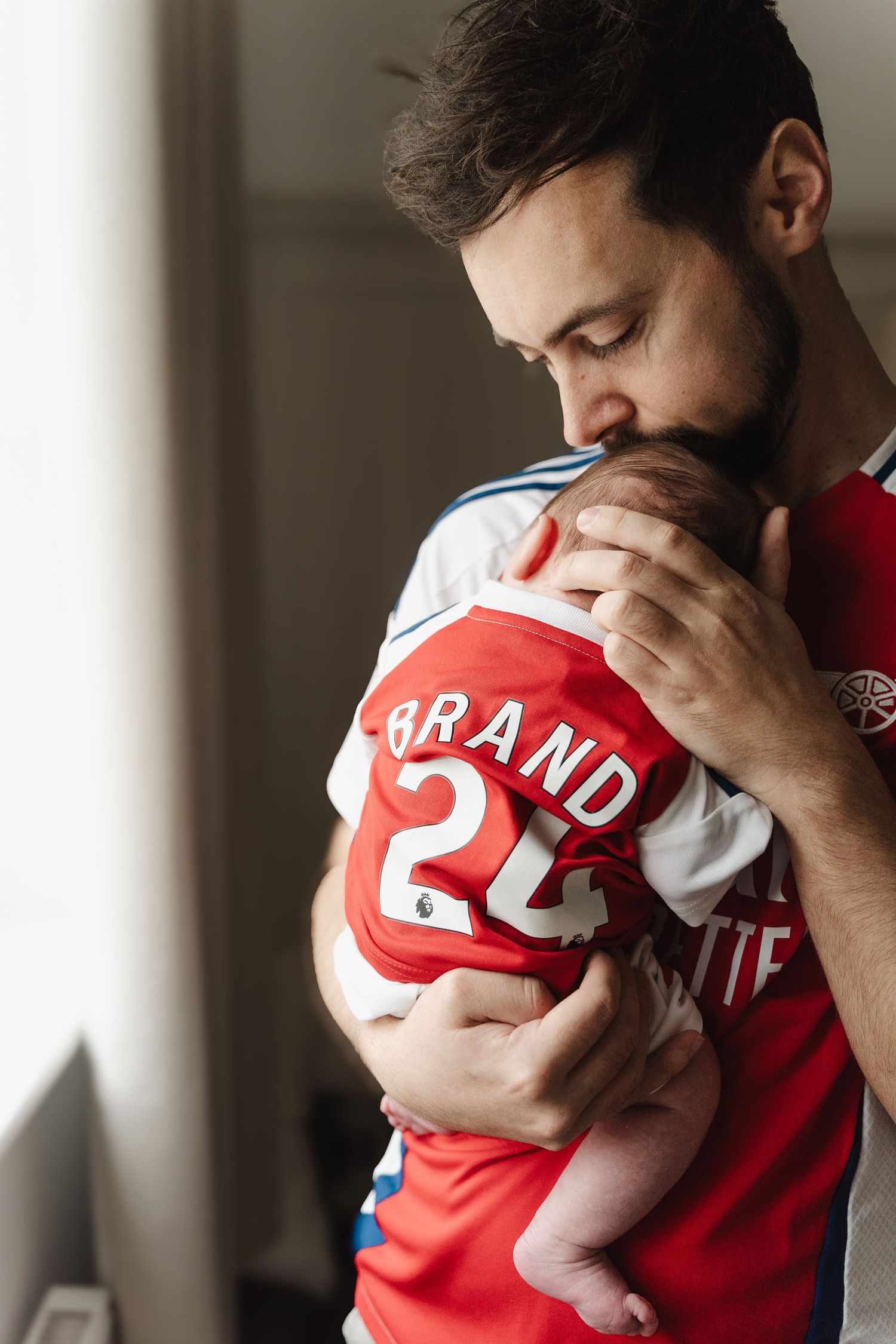 A parent in a red sports jersey tenderly holds and kisses a sleeping baby in matching team attire.