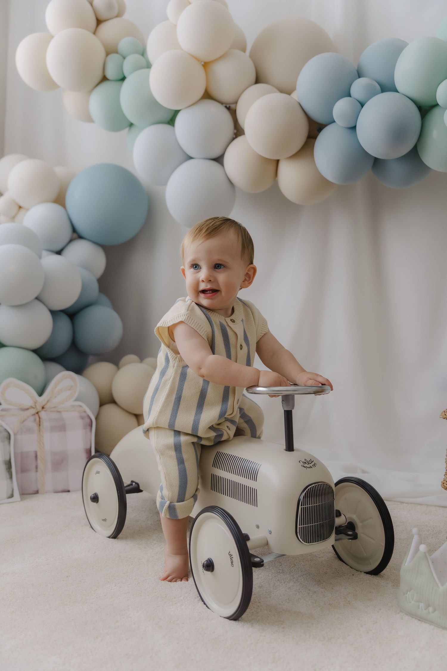 Baby riding vintage-style white ride-on car with blue and white balloon arch backdrop.