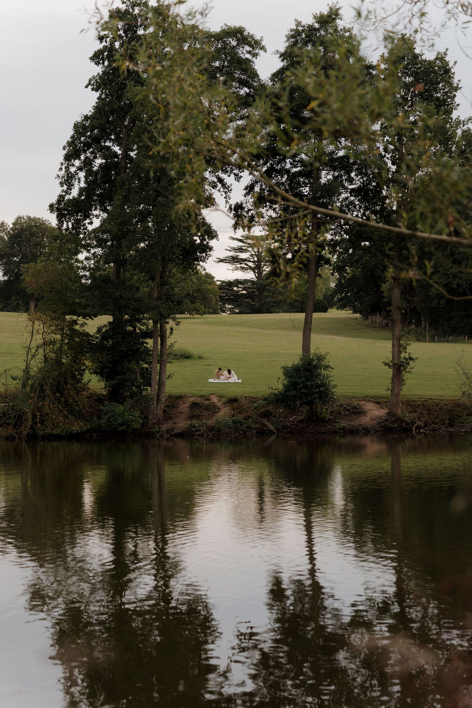 A serene lake reflects trees along its banks under cloudy skies during autumn.