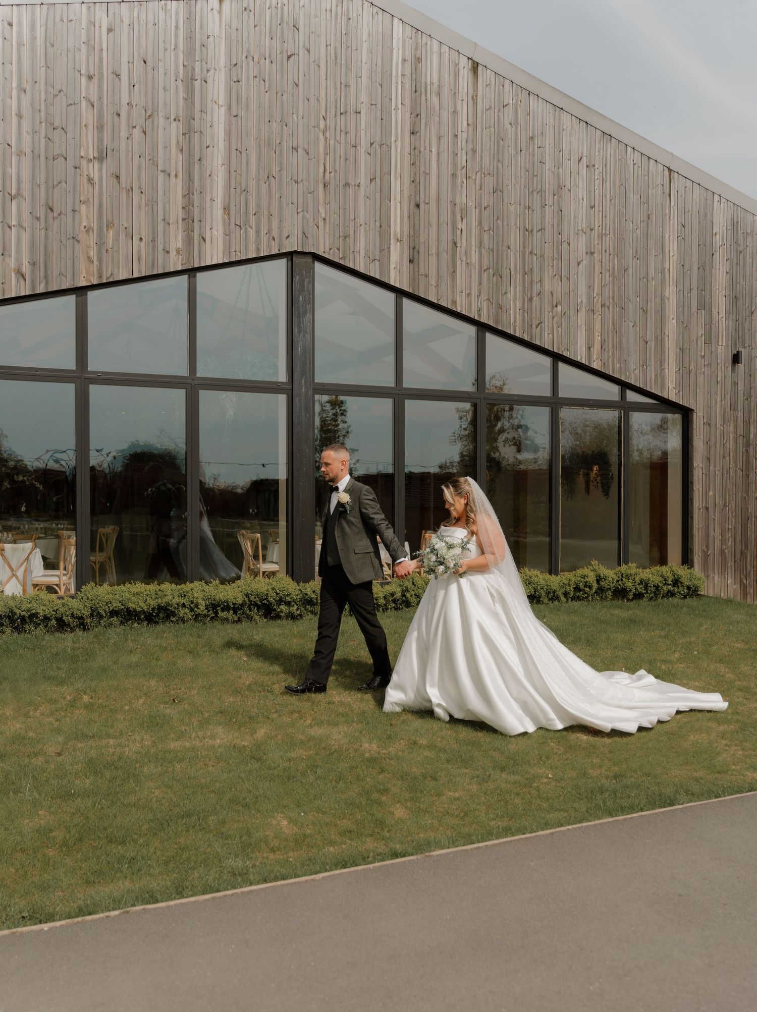 Bride and groom walk hand in hand on green lawn outside modern wooden wedding venue with large glass windows.