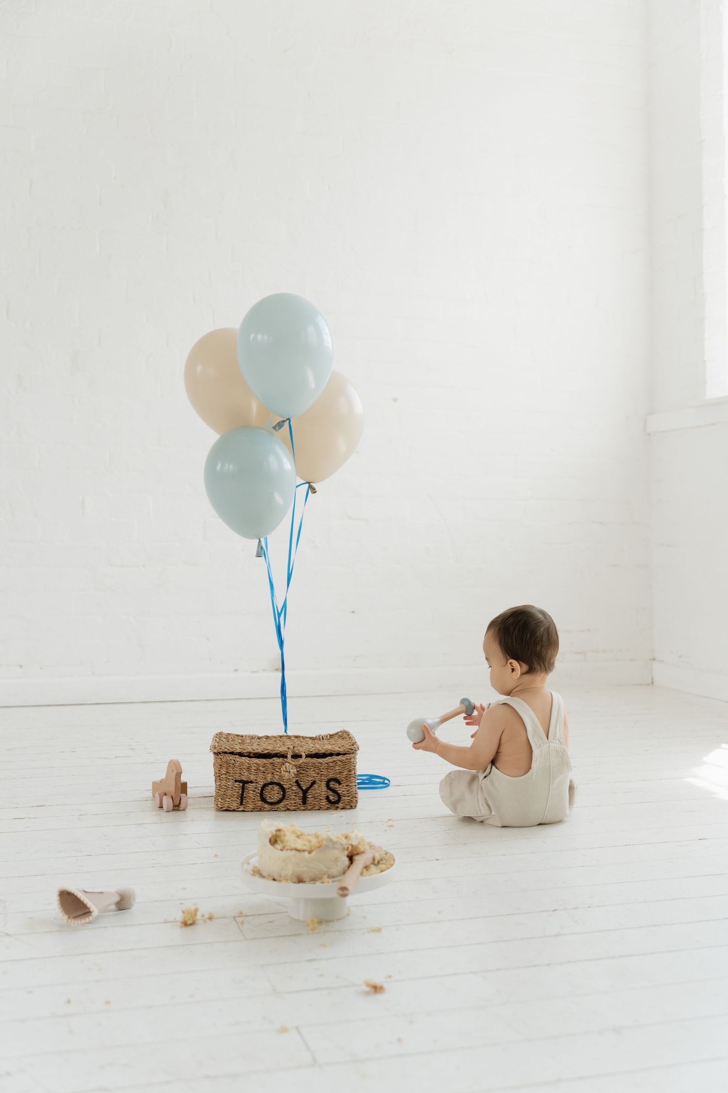 A baby sits on a white floor near a wicker toy basket with blue and cream balloons, surrounded by smashed birthday cake.