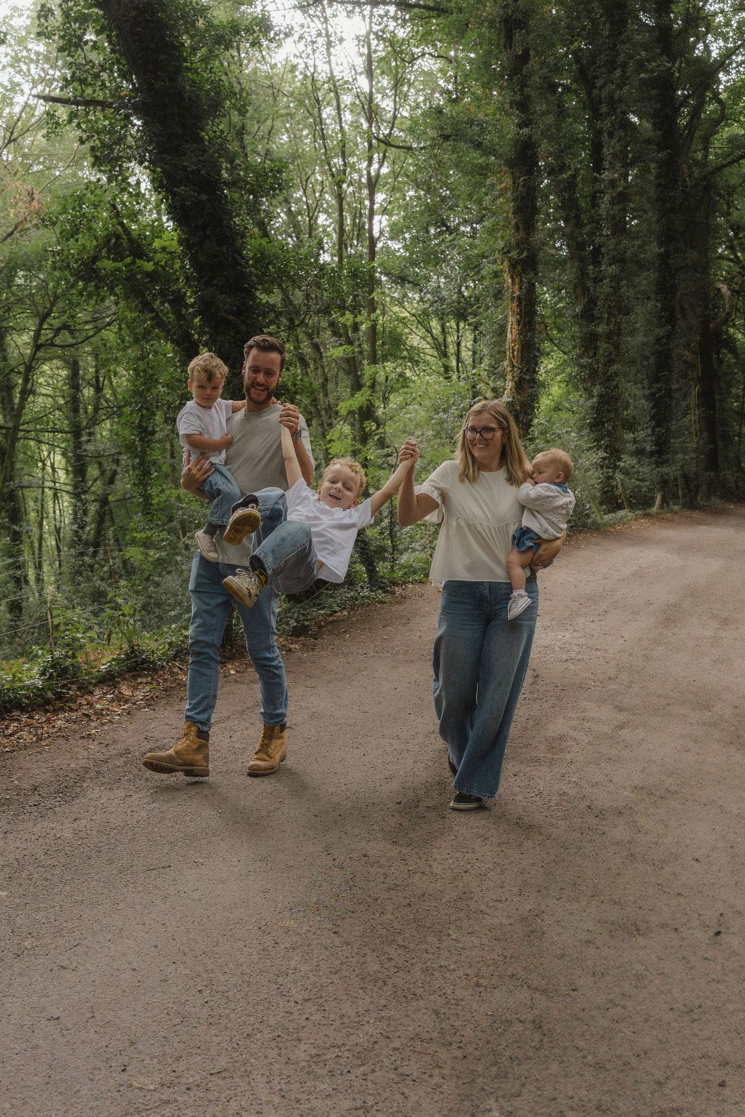 A family enjoys a playful moment together on a dirt path surrounded by lush green trees on a sunny day. Derbyshire outdoor photography.