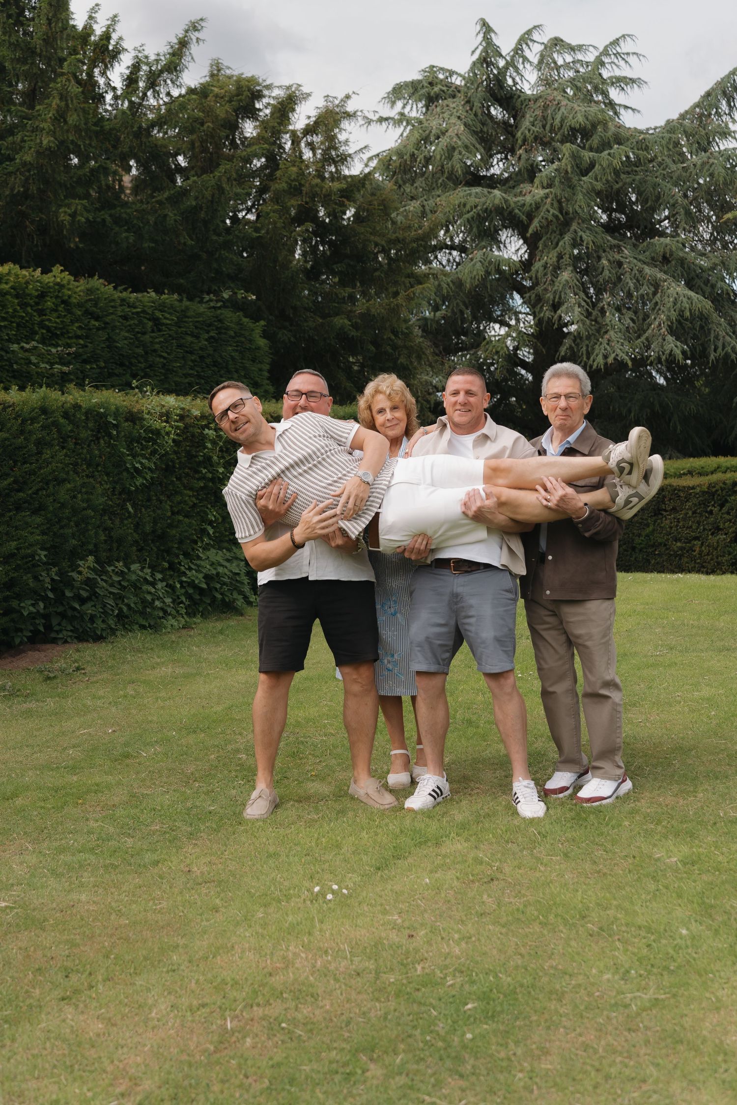 Group of five adults posing outdoors on grass, two men carrying others, with green hedges and trees in background.