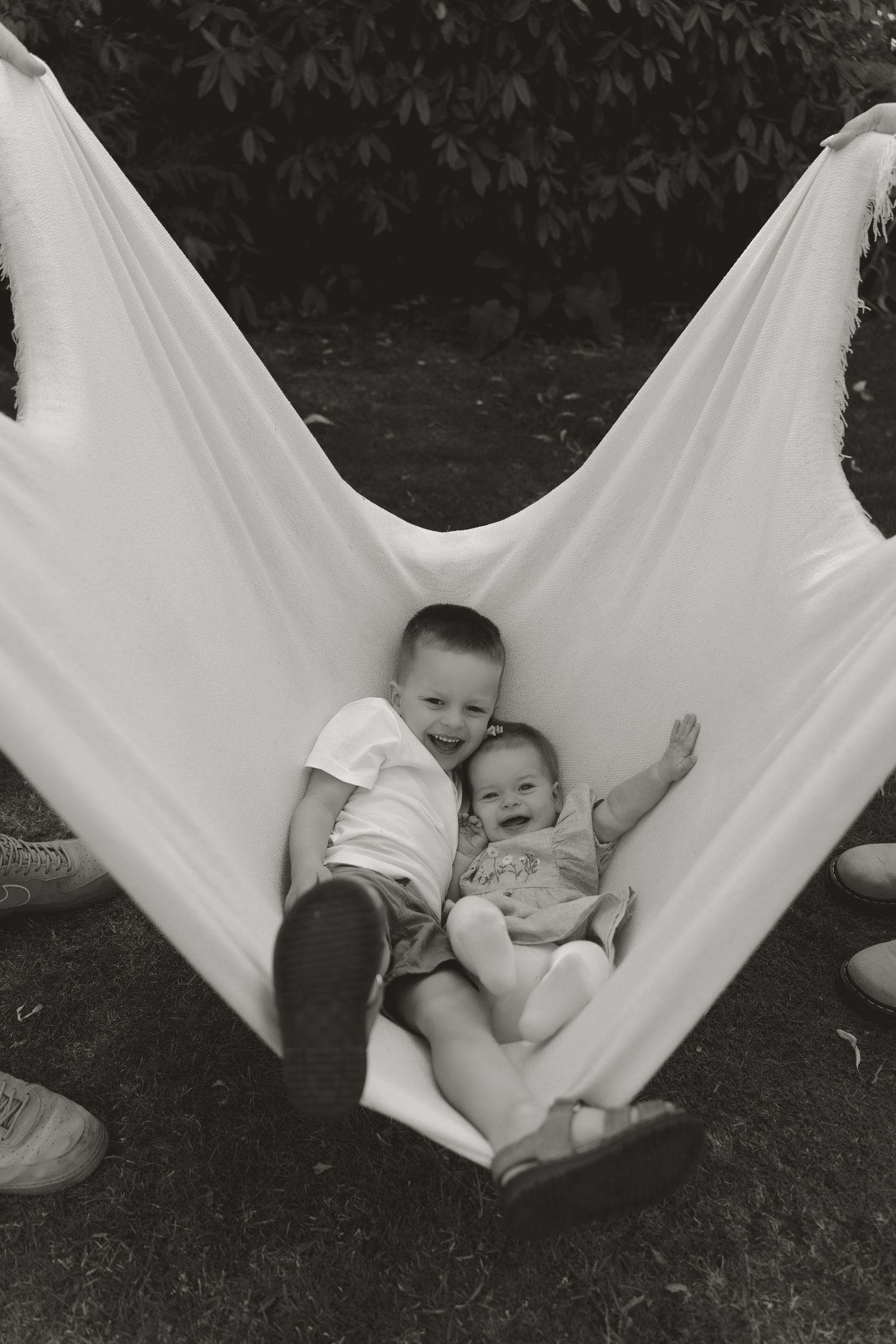 Two young children laughing and relaxing together in a white fabric hammock outdoors, black and white photo.