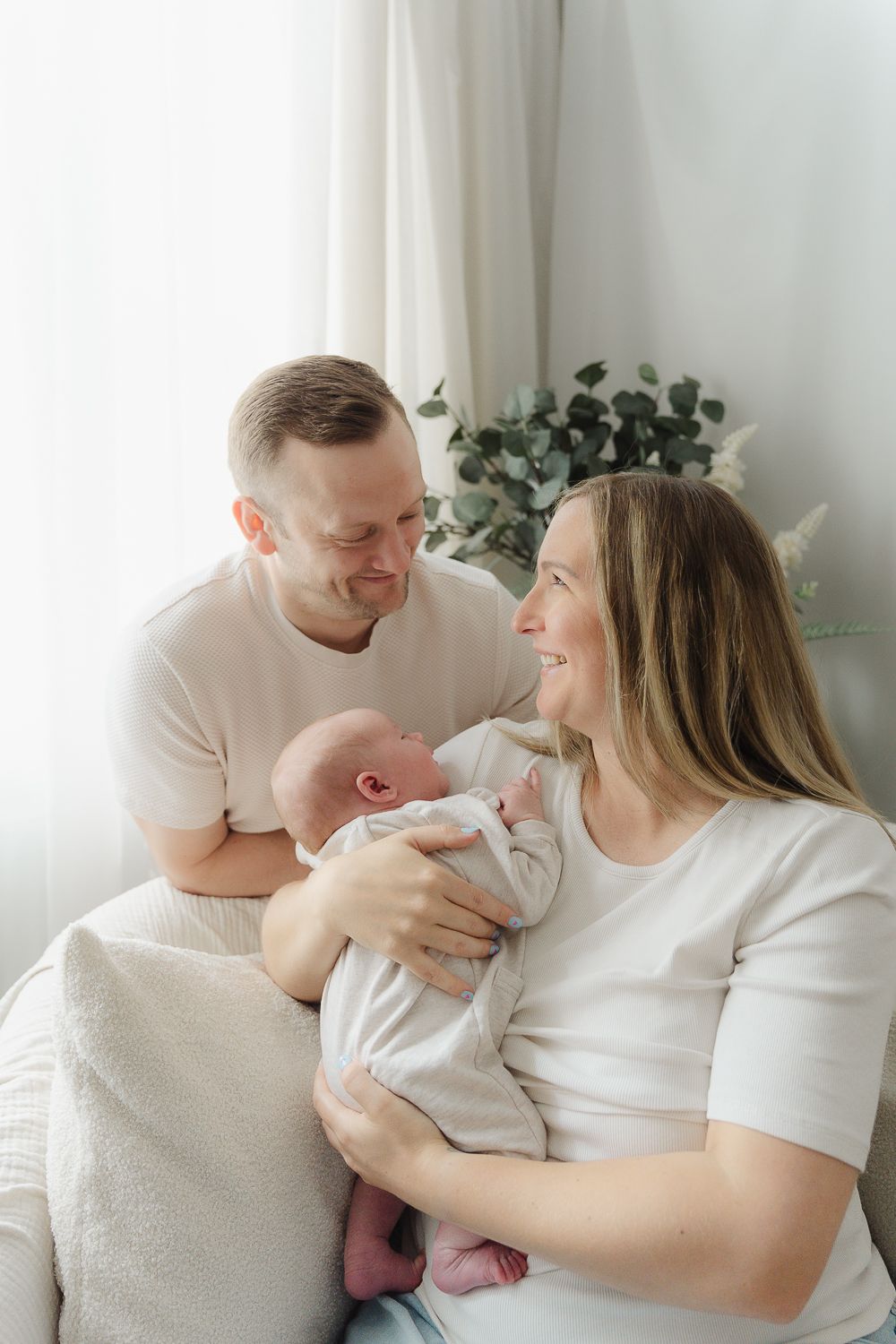 A couple in white shirts shares tender moments with their newborn baby against a light backdrop.