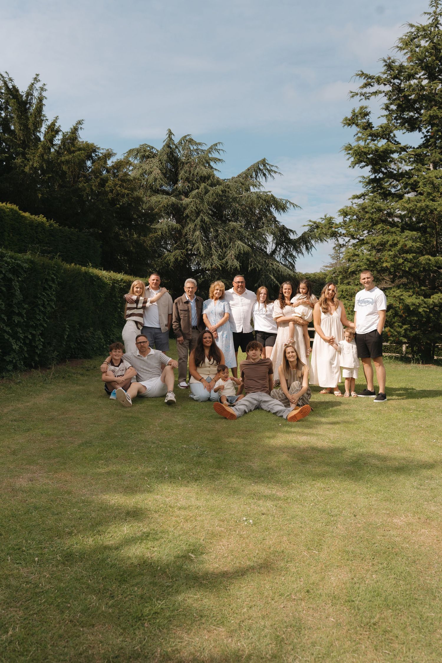 Large family group photo outdoors on green lawn surrounded by tall trees and hedges on a sunny day.