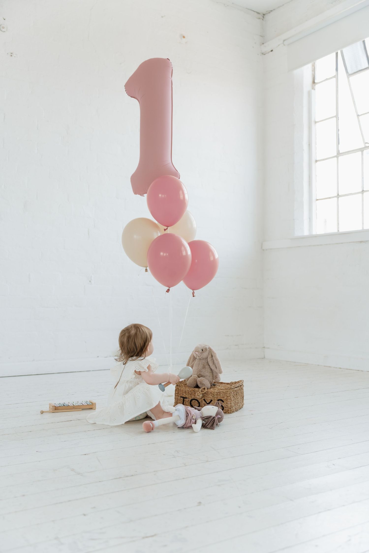 A baby girl sits on a white floor with a bunny toy and pink balloons celebrating her 1st birthday.