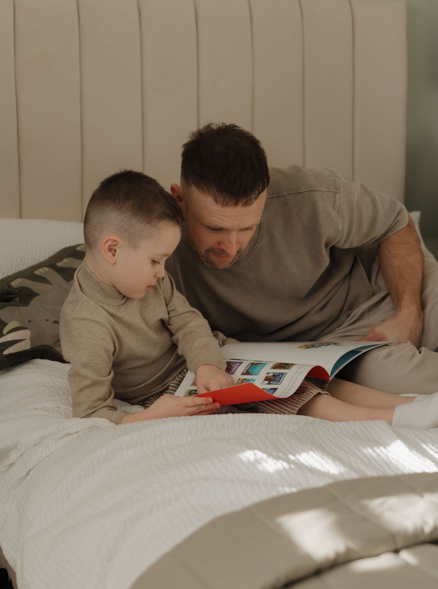 A father and young son sit together on a bed, reading a colorful book in a cozy, sunlit bedroom.