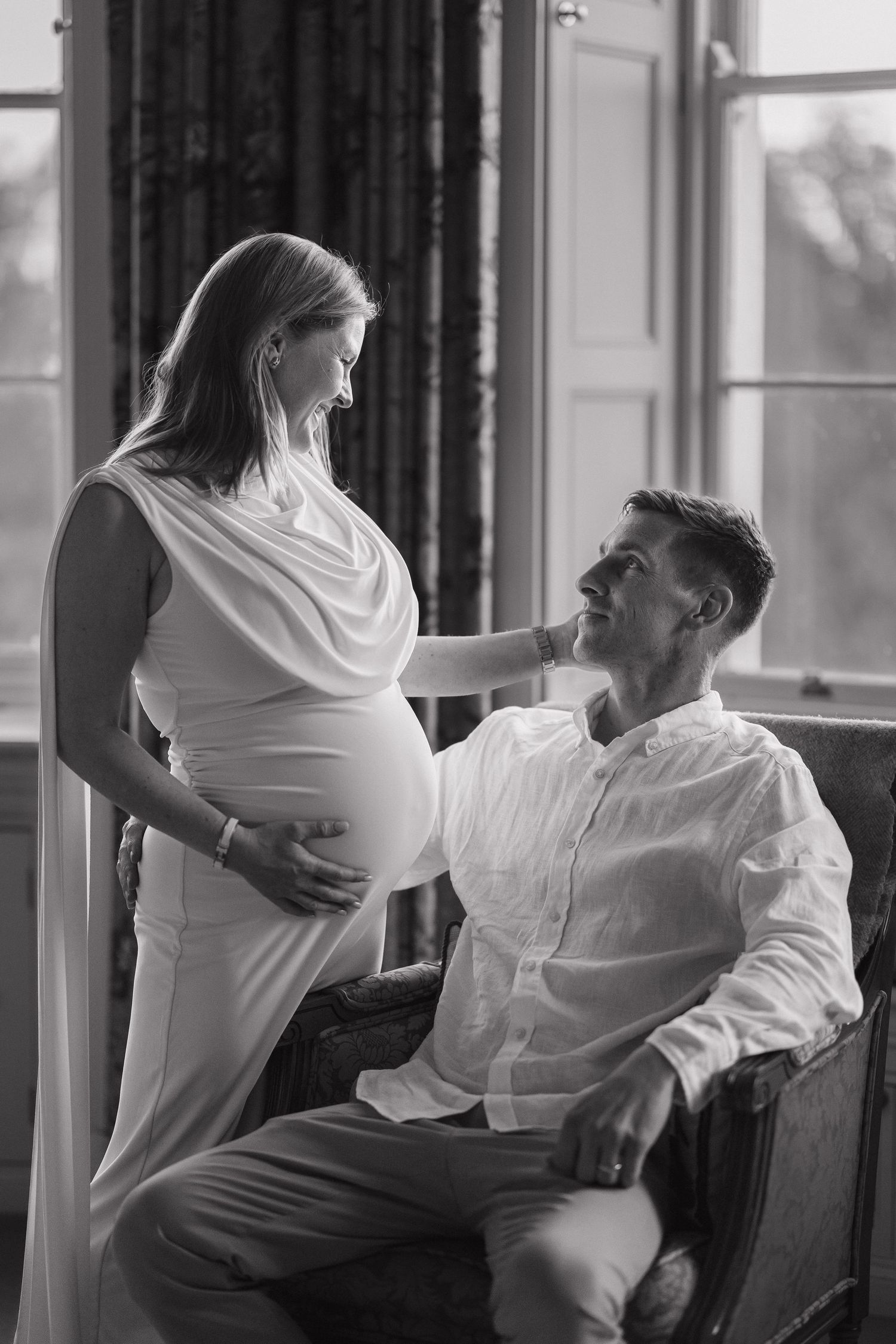 Black and white photo of a pregnant woman in white dress with her partner seated, sharing a tender moment by a window.