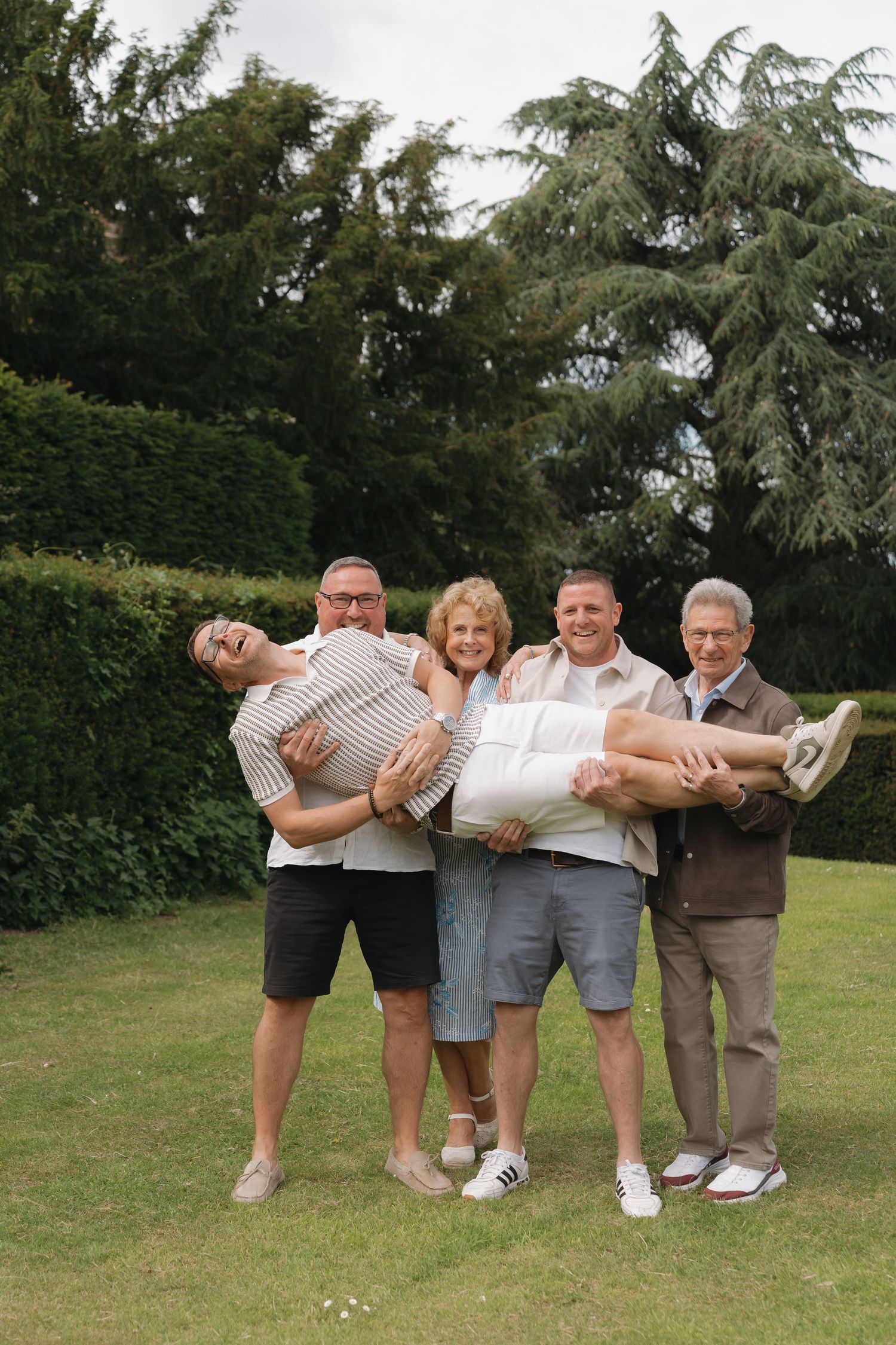 Group of five adults posing outdoors, two men carrying a woman horizontally while others smile in a garden setting.