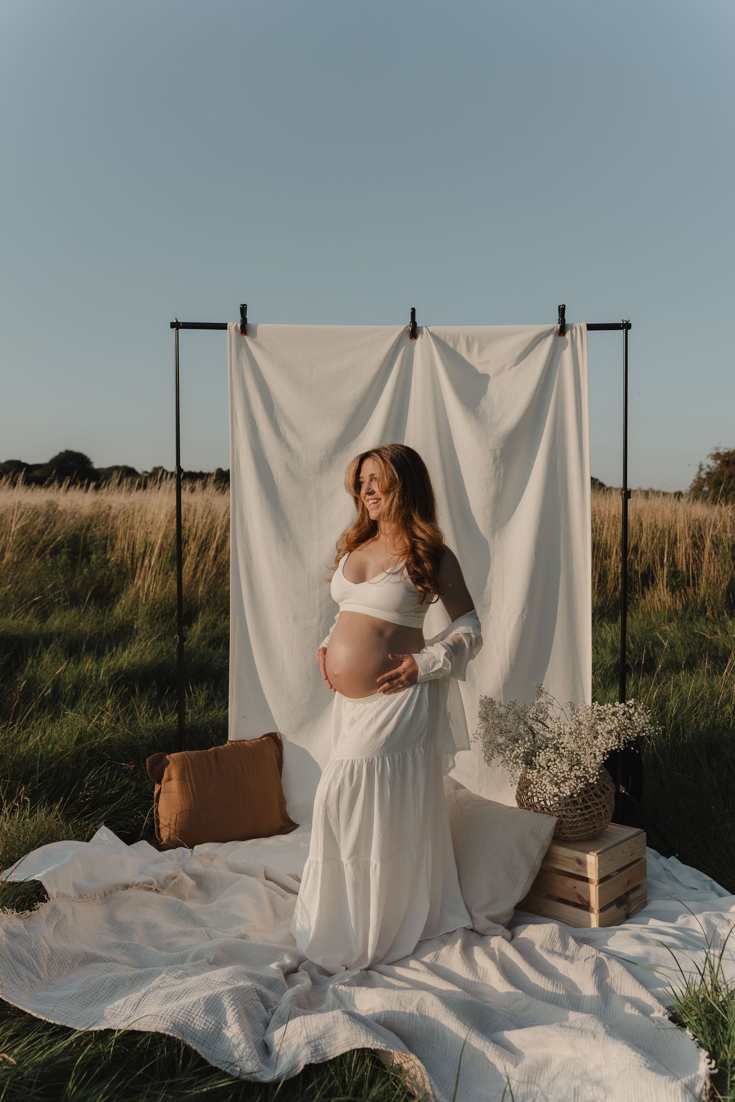 Ethereal maternity photoshoot in a field with white backdrop, flowing dress at golden hour. Taken by Shots by Maz in Derbyshire