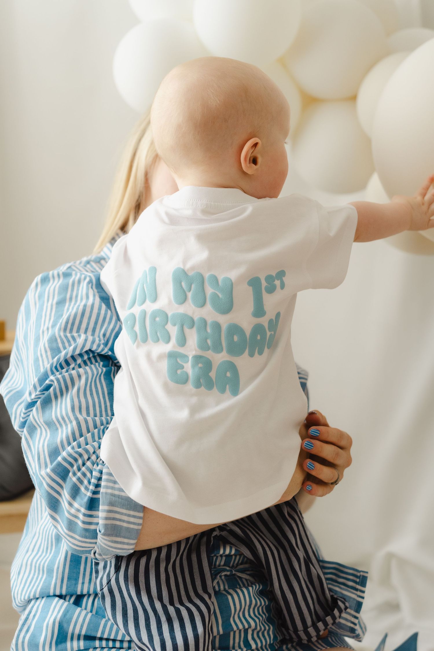 Baby wearing white and blue striped outfit crawls while looking up at white balloon decorations.