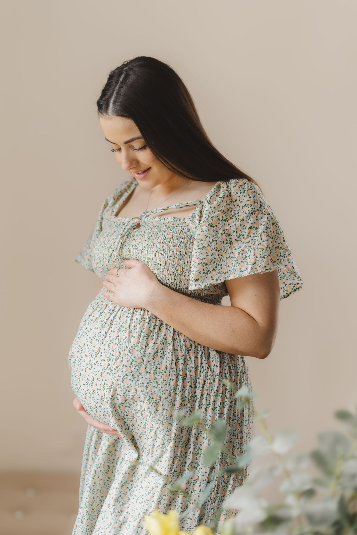 A pregnant woman in a floral dress touches her belly while posing against a neutral beige background.