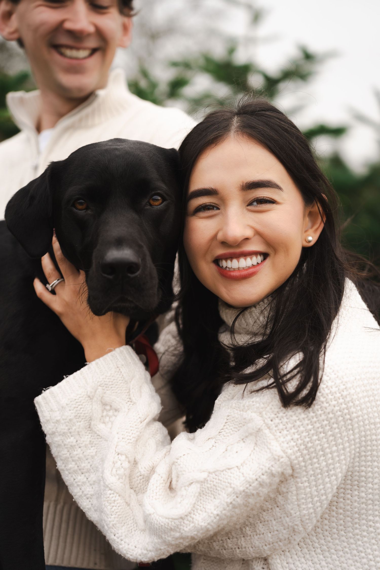 A beautiful black Labrador dog poses with people wearing cozy white sweaters outdoors.