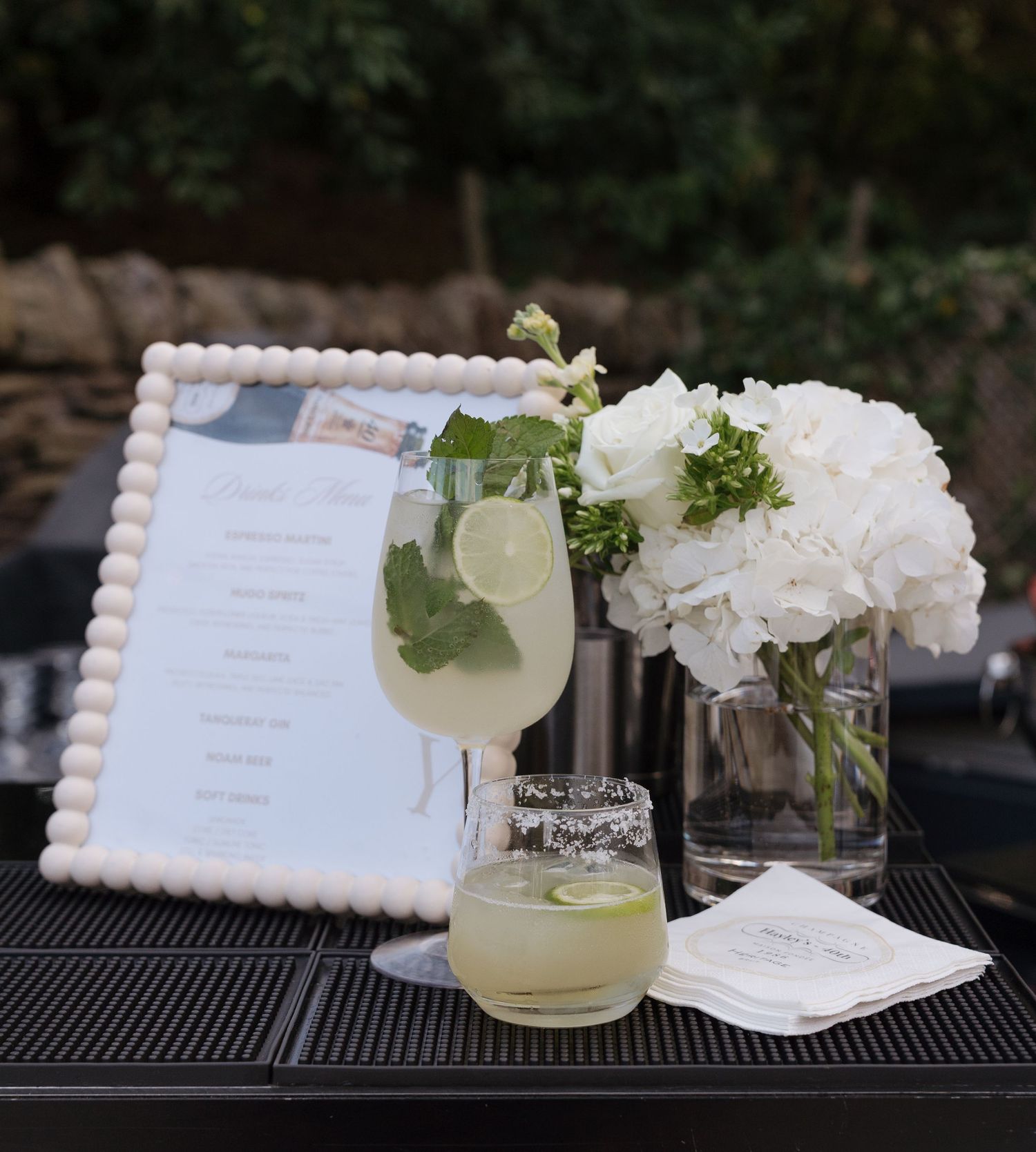 Elegant cocktail bar setup with white hydrangeas, framed drink menu, and garnished cocktails on a black surface.