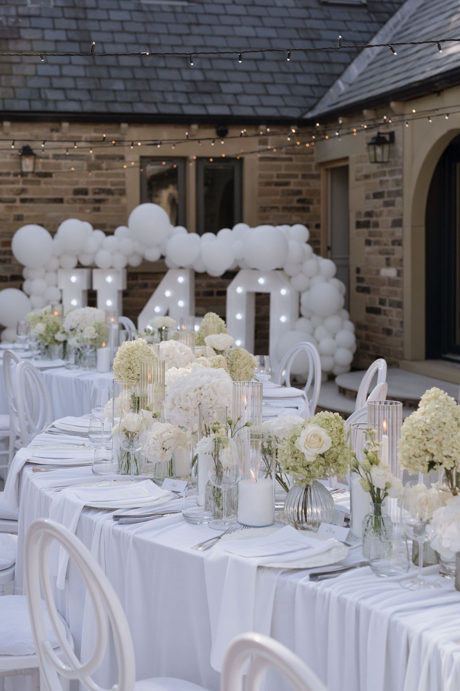 Elegant all-white outdoor wedding reception with floral centerpieces, balloon arch, marquee letters, and string lights.