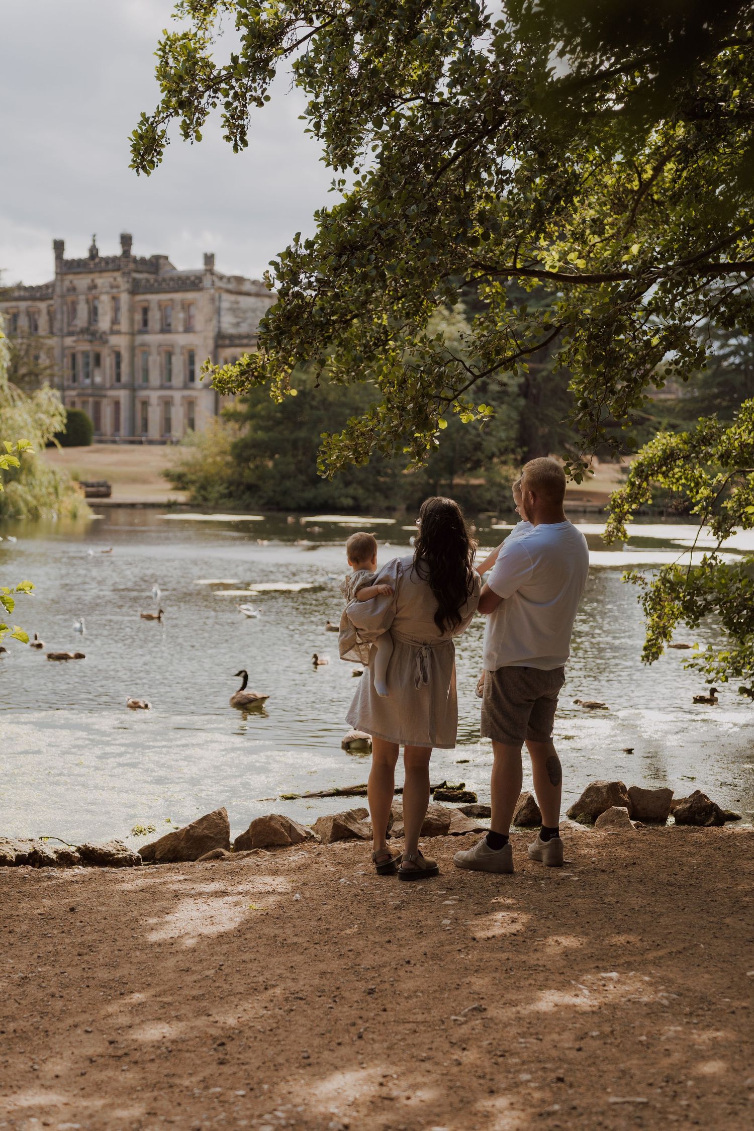 Family standing by a serene lake with ducks, admiring a grand historic mansion surrounded by lush trees.