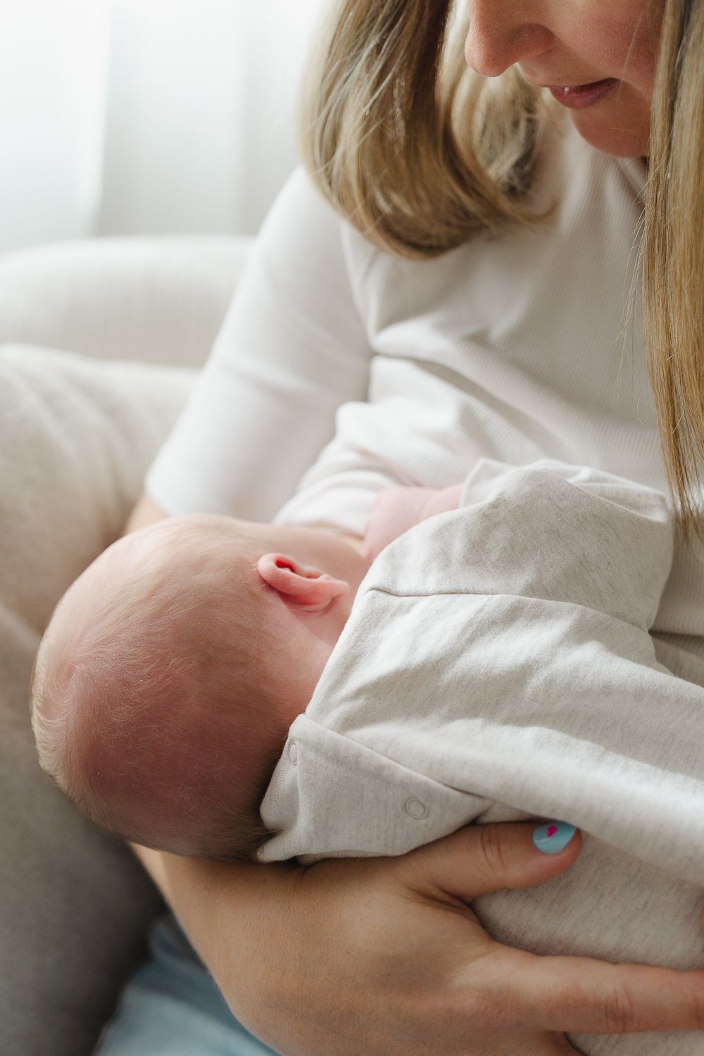 A parent in a white sweater cradles and nurses a newborn baby wrapped in a soft white blanket.
