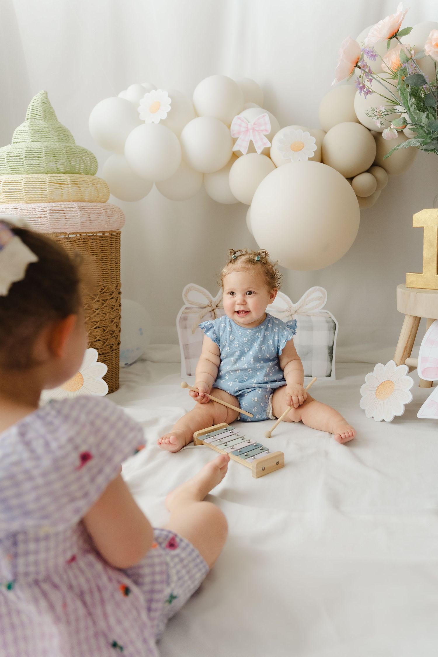 Dreamy first birthday cake smash photo session with white balloons, floral decorations and a blue romper against white backdrop.