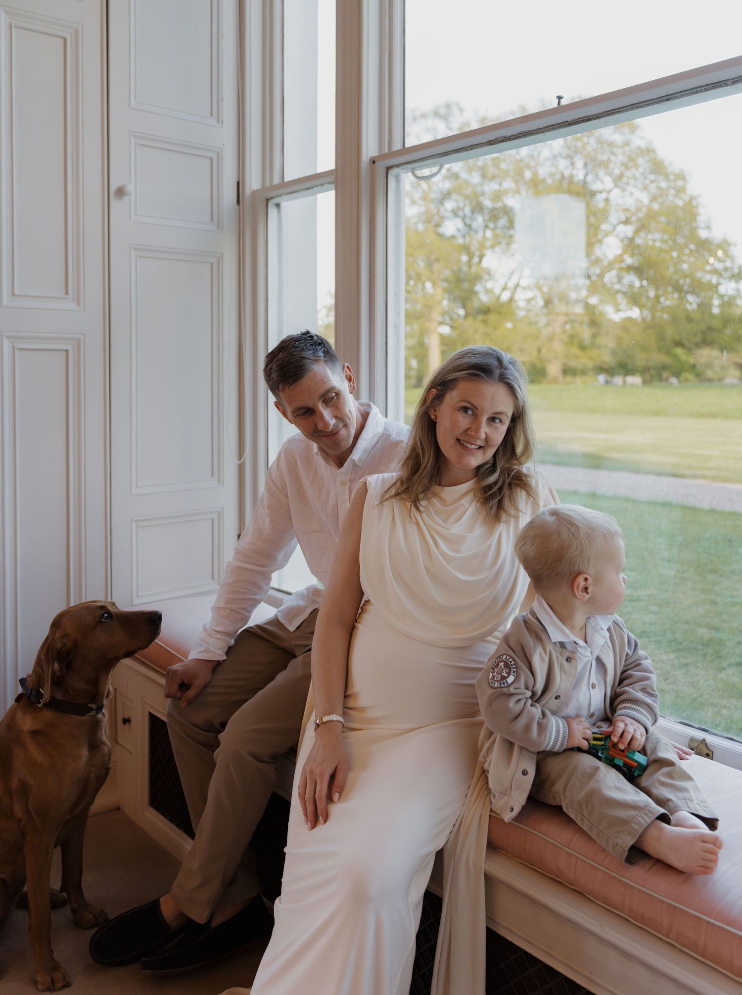 A couple with a toddler and brown dog pose by a large window in an elegant white room with garden views.