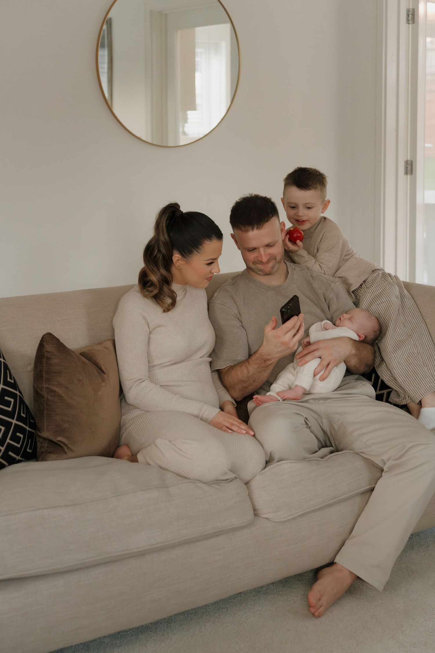 Family of four sitting on a beige sofa, parents admiring newborn baby while young boy watches over their shoulders.