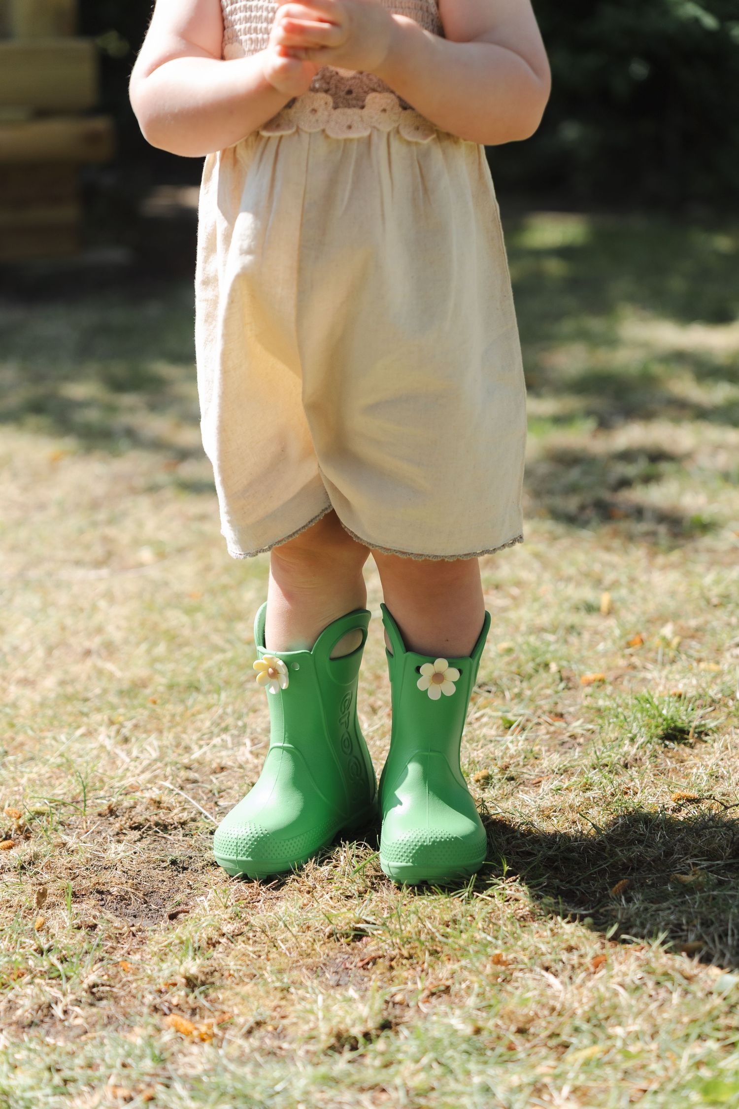 Toddler wearing green rubber rain boots with flower details and a cream romper standing on grass.