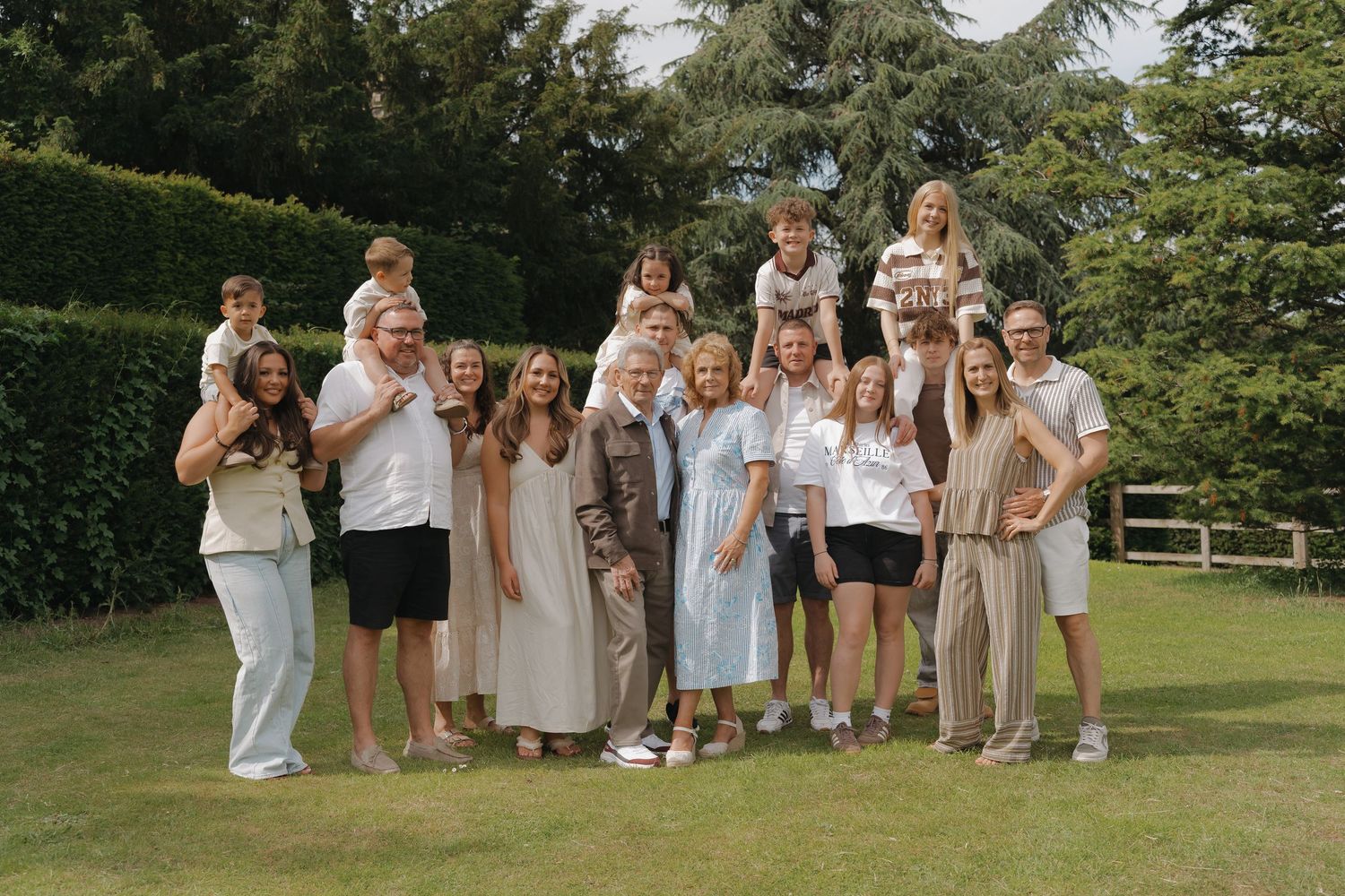 Large family group posing together on green lawn surrounded by lush hedges and trees on a sunny day.