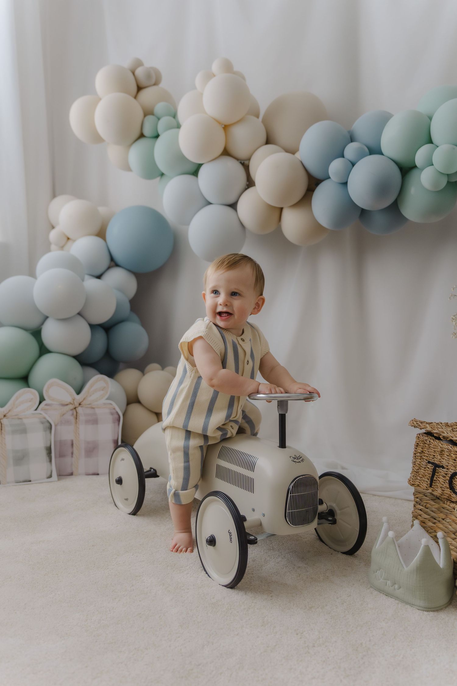 A baby rides a vintage-style white metal ride-on car against a backdrop of blue and cream balloon garland decorations.