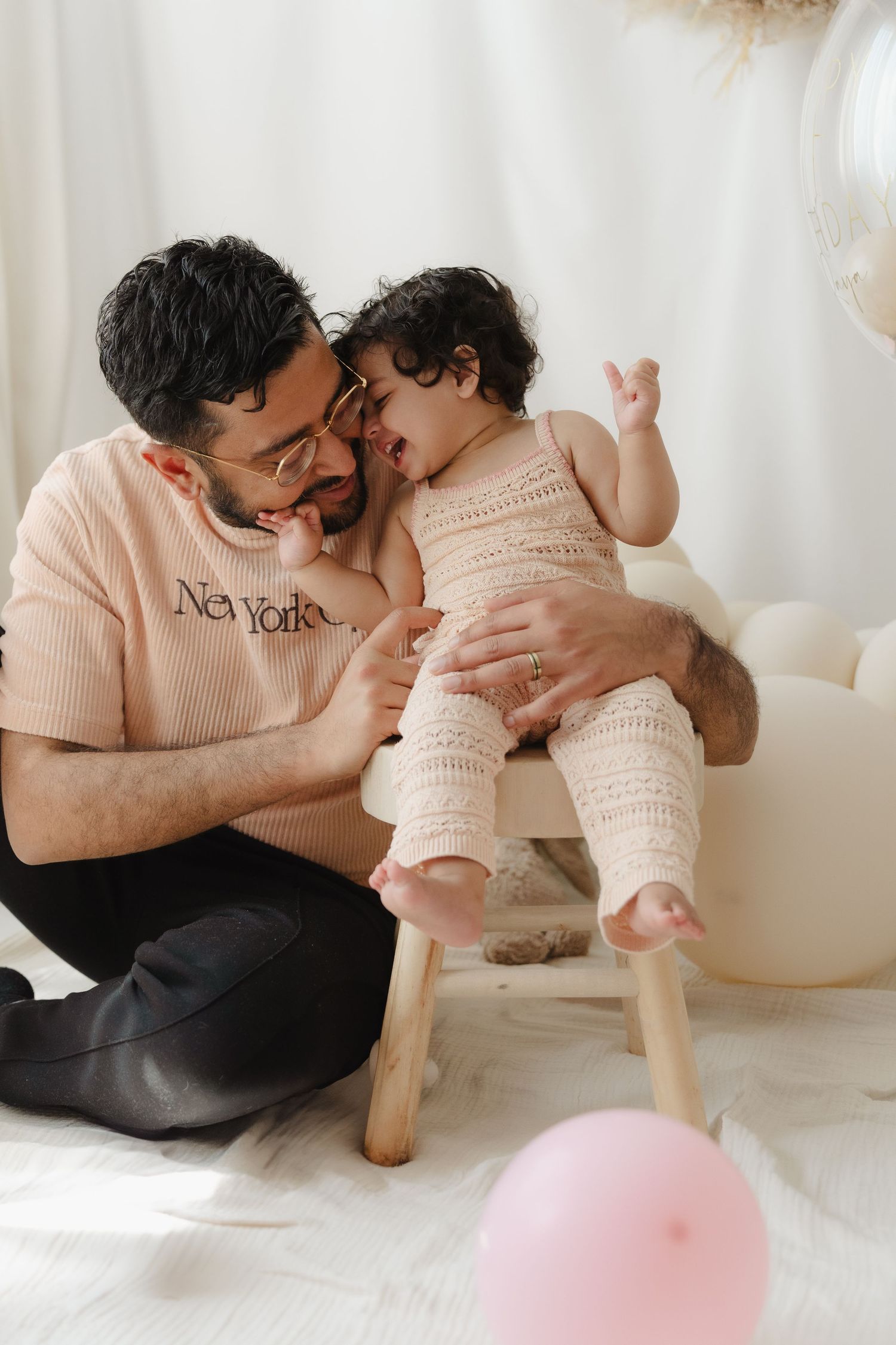 Parent and baby in matching pink sweaters share tender moments while sitting on white floor with pink balloons.