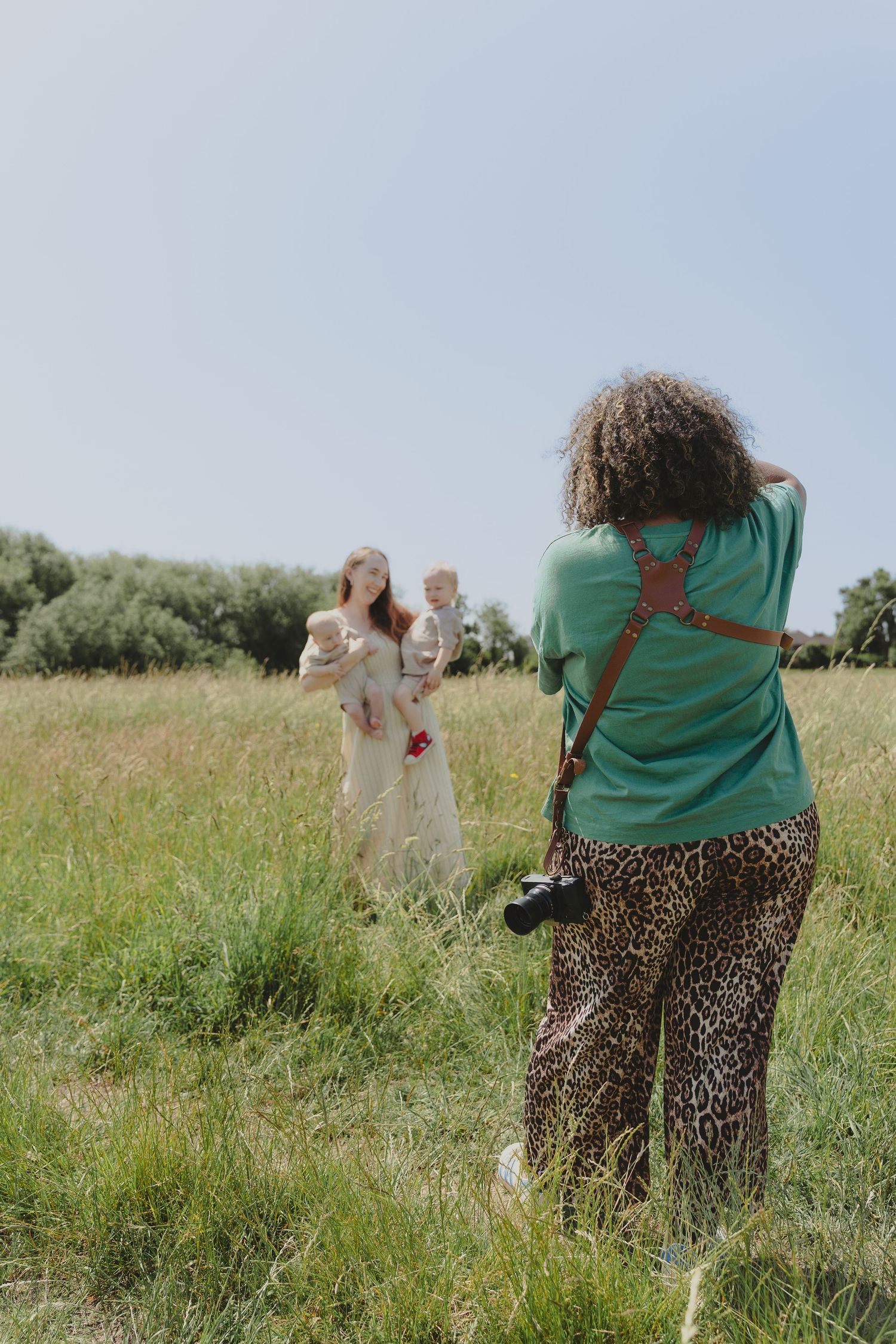 A photographer wearing a green top and leopard print pants takes pictures in a grassy field on a bright summer day.