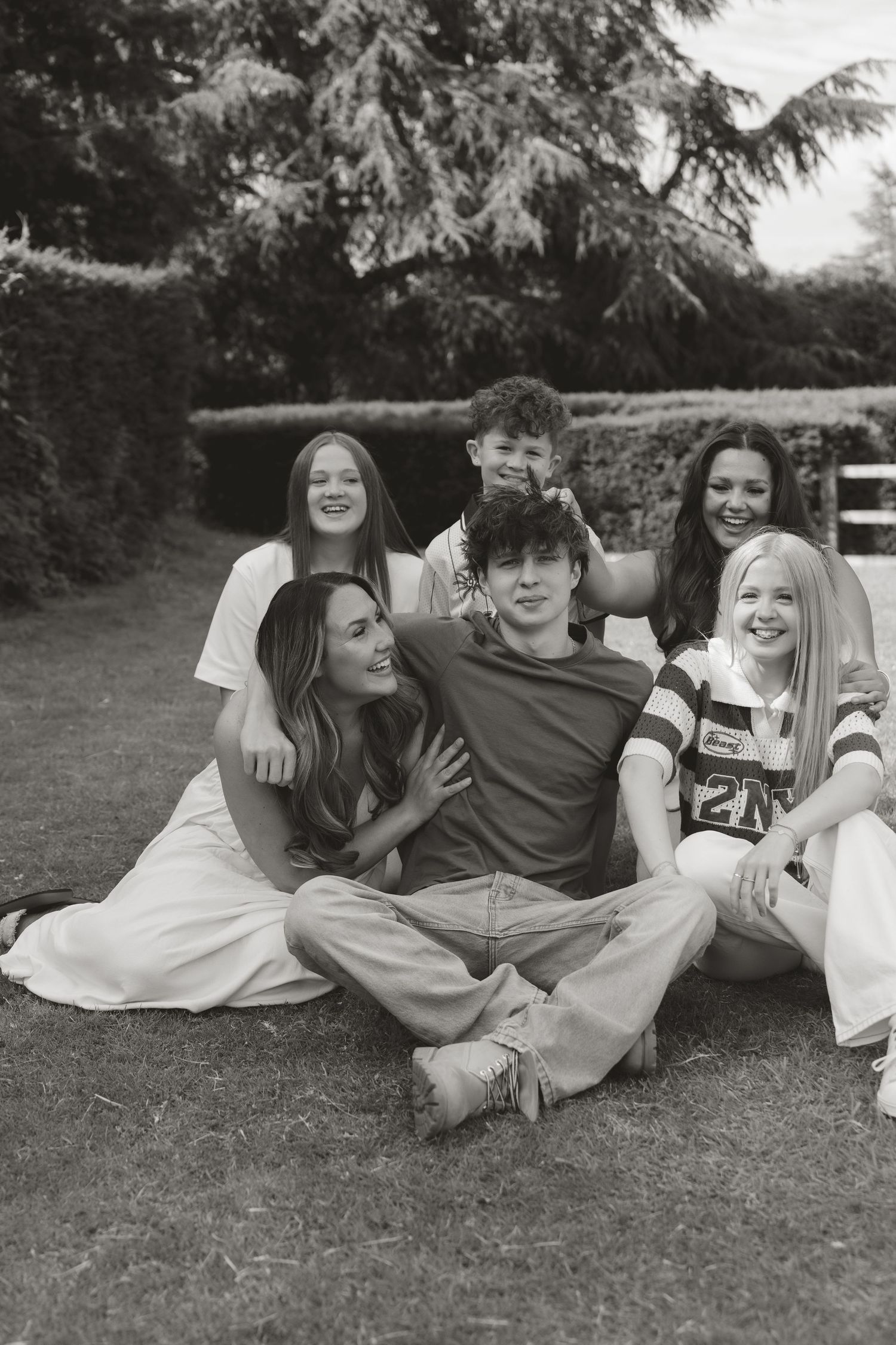 Black and white photo of six young people smiling and posing together on a lawn surrounded by hedges.